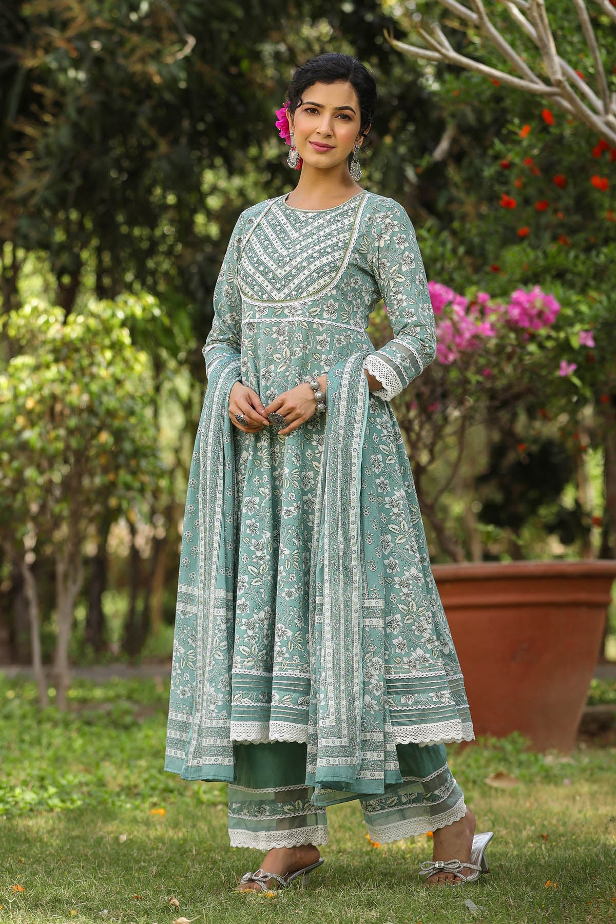 Woman in a green traditional outfit standing outdoors with trees and flowers in the background