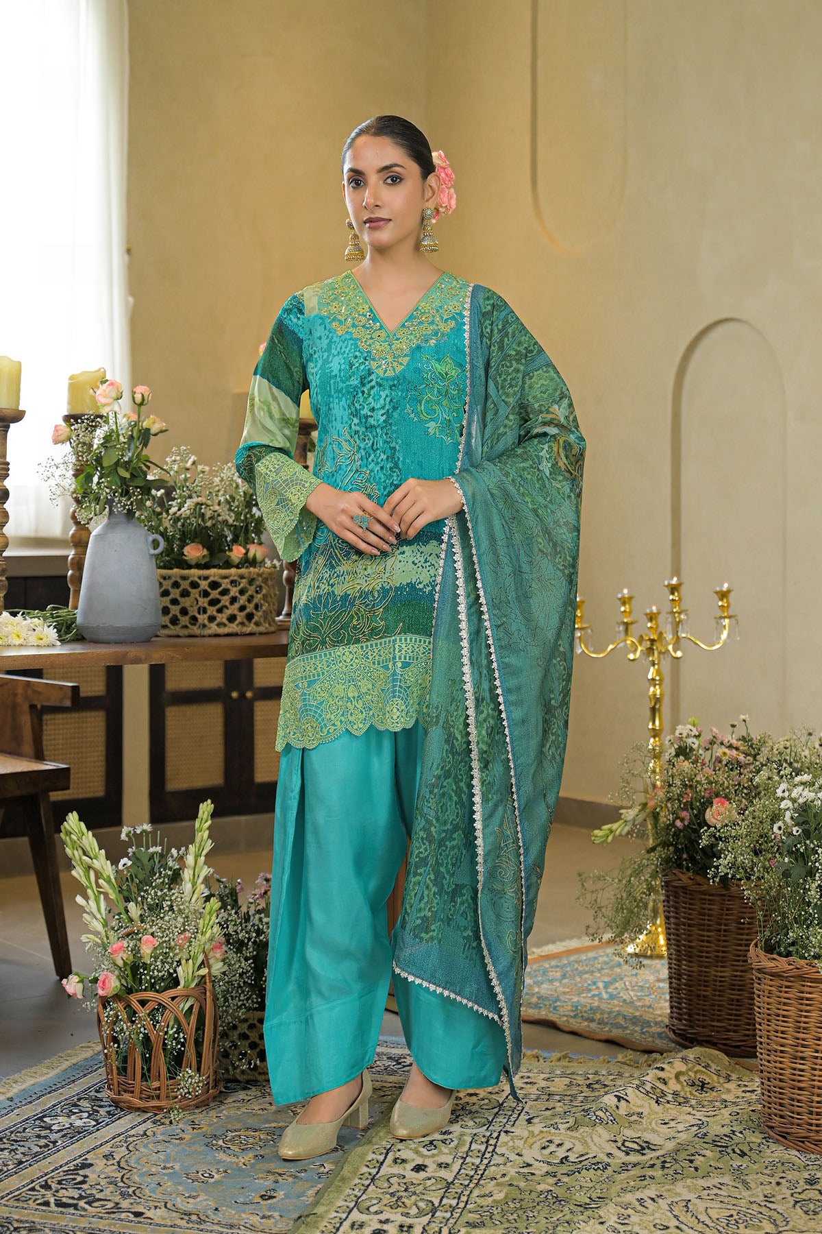 Woman in a teal traditional outfit standing in an elegant room with floral decorations.