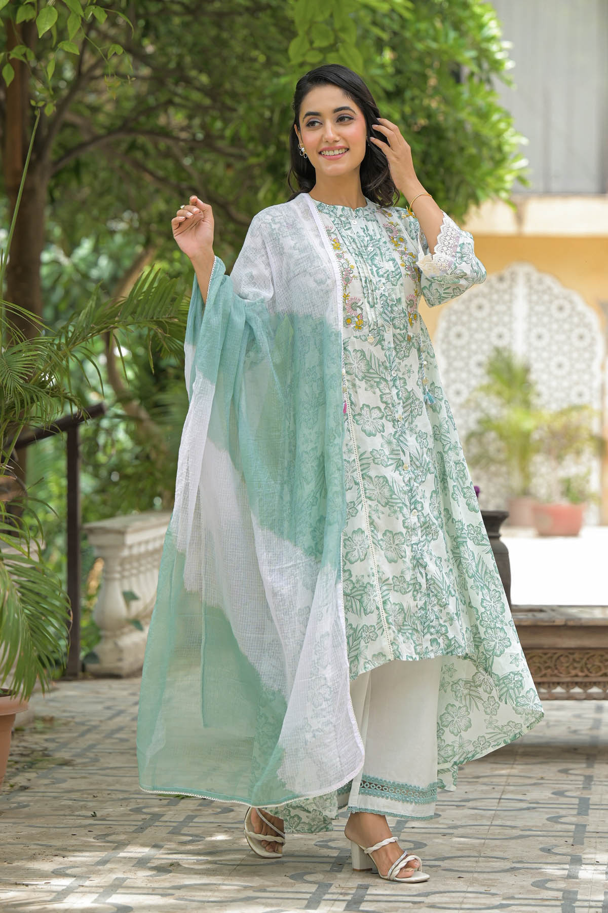 Woman in a light green and white traditional outfit standing outdoors with greenery in the background
