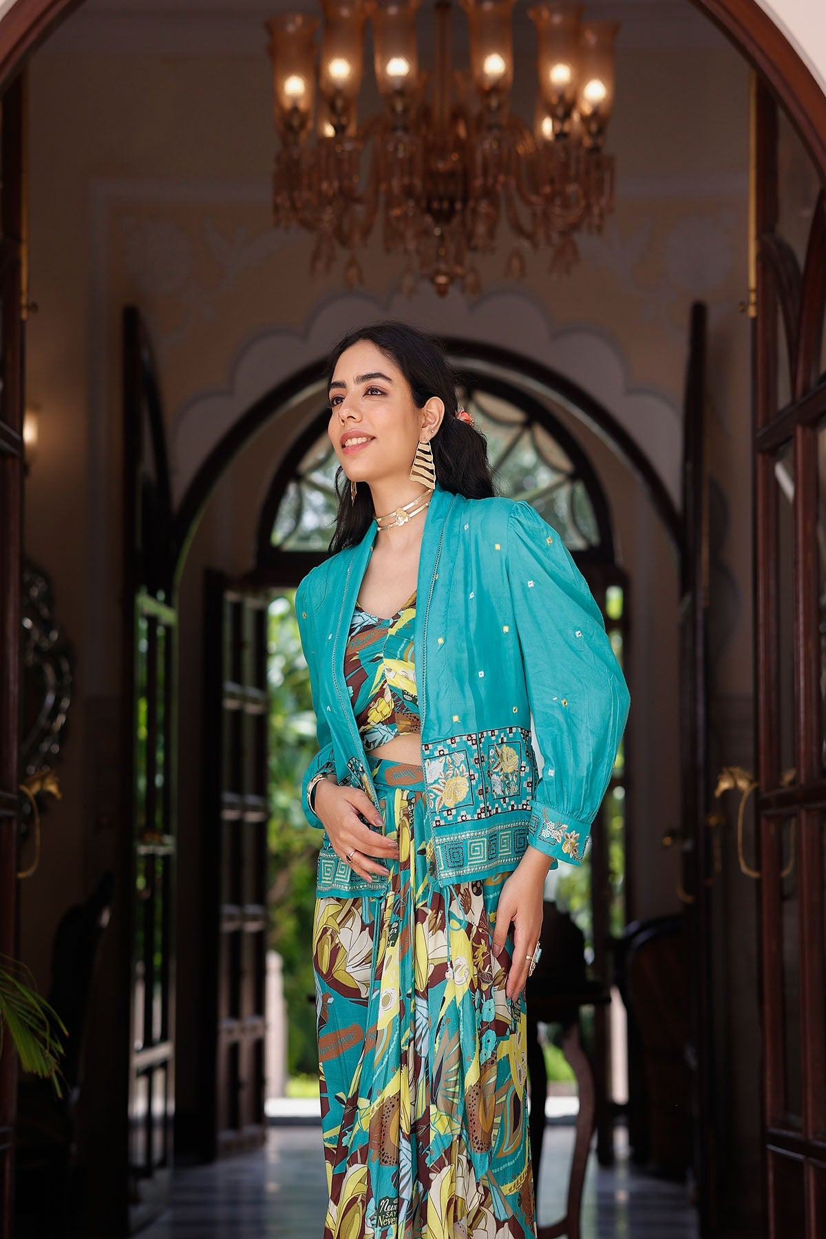 Woman in a colorful traditional outfit standing in an elegant interior setting with a chandelier.