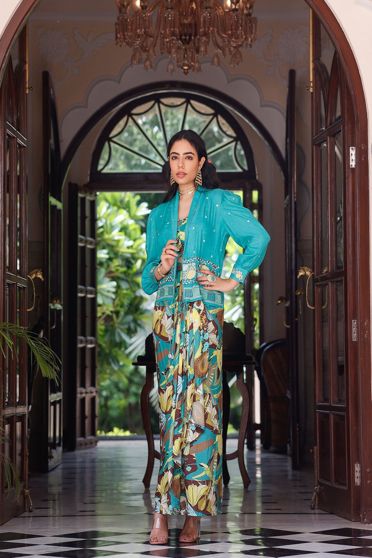 Woman in a turquoise outfit standing in an elegant hallway with a chandelier.