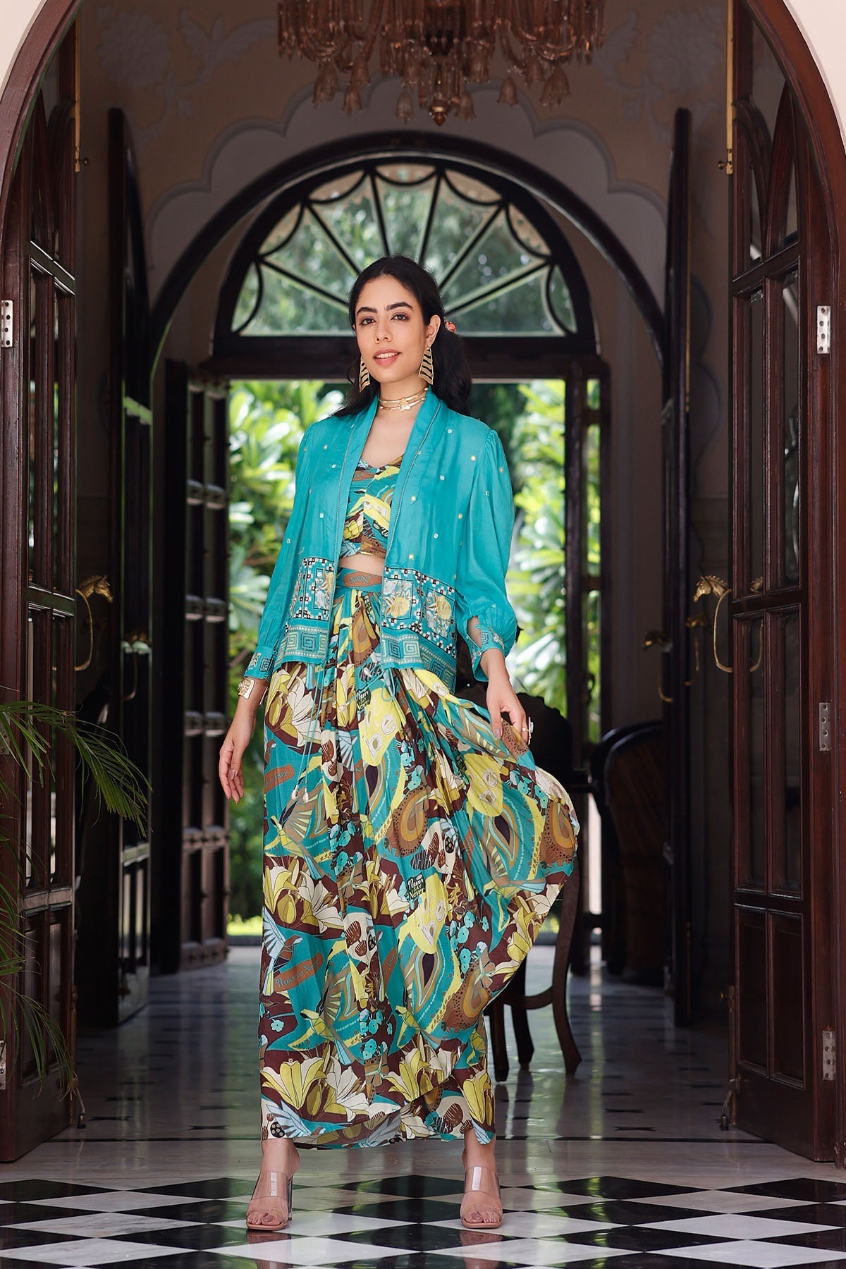 Woman in a floral dress and turquoise jacket standing in a doorway with a decorative ceiling.