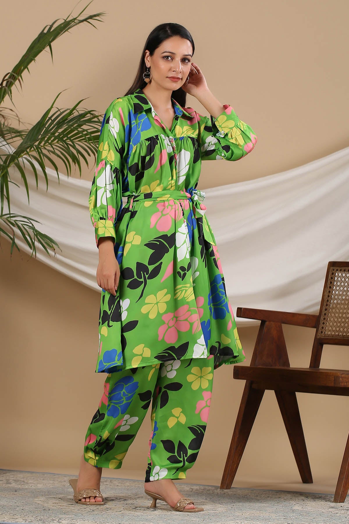 Woman wearing a green floral outfit standing against a beige wall with a wooden chair and plant.