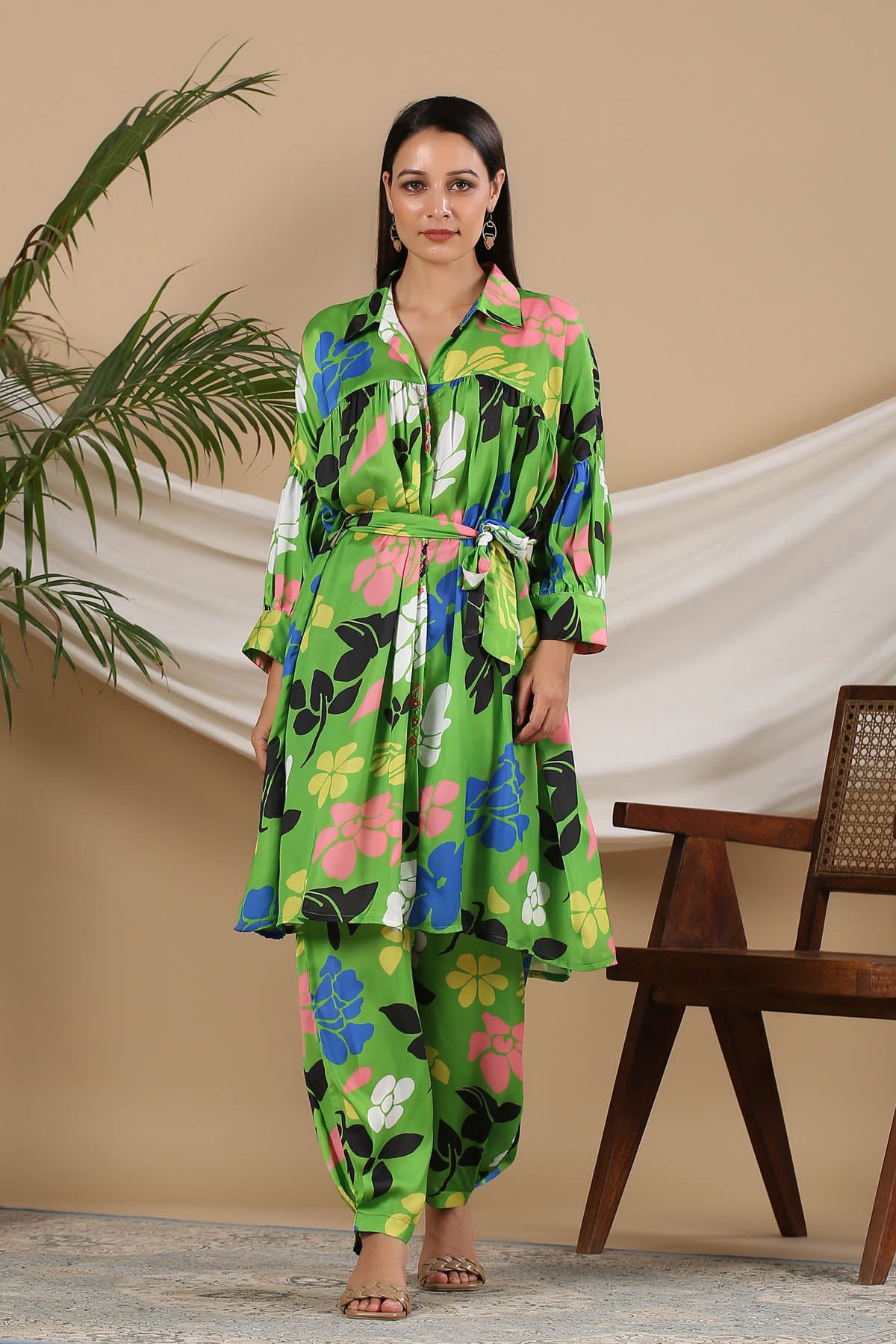 Woman wearing a colorful floral outfit standing in a room with a plant and wooden chair.