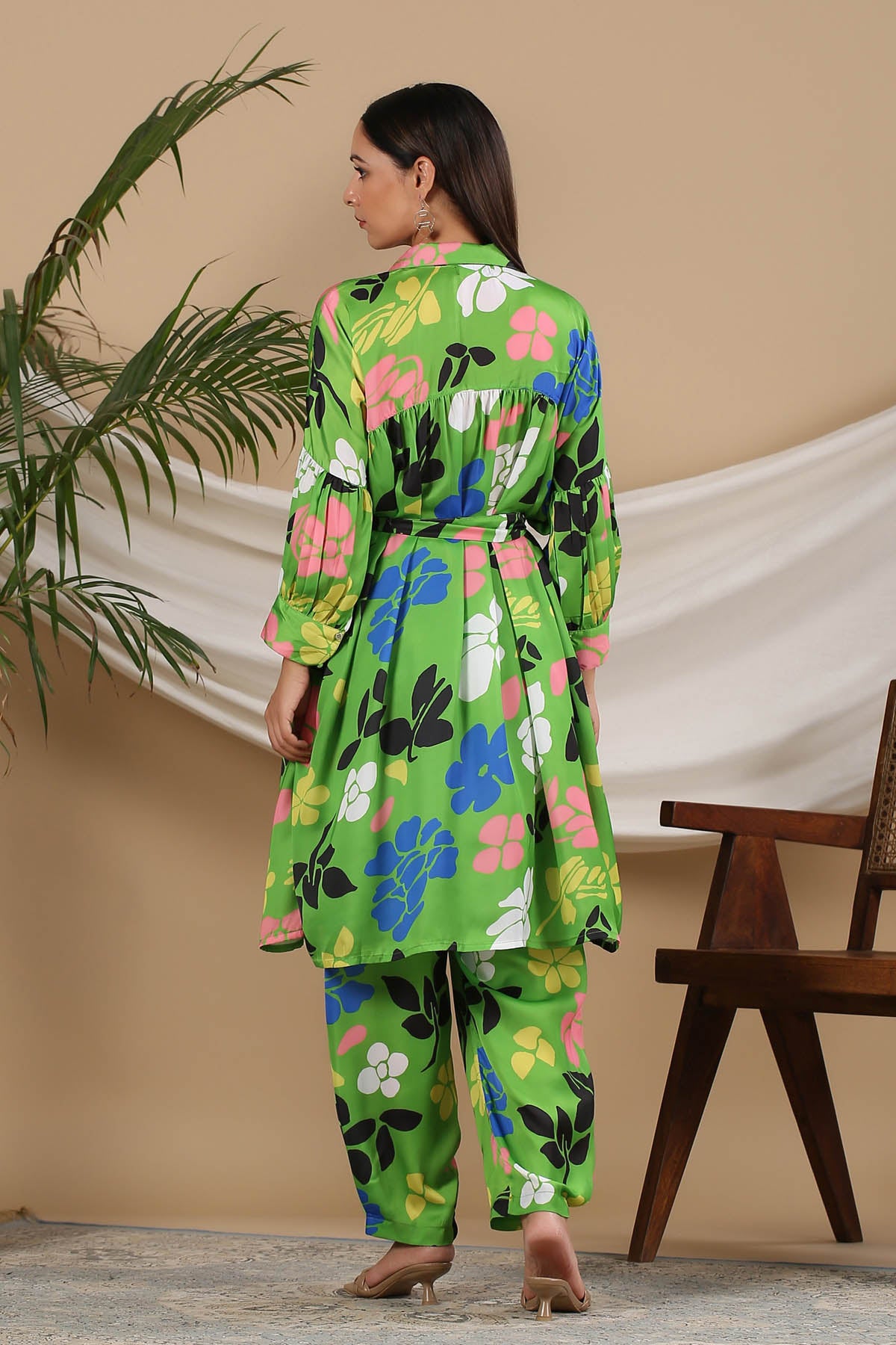 Woman wearing a green floral outfit standing in a room with a plant and chair.