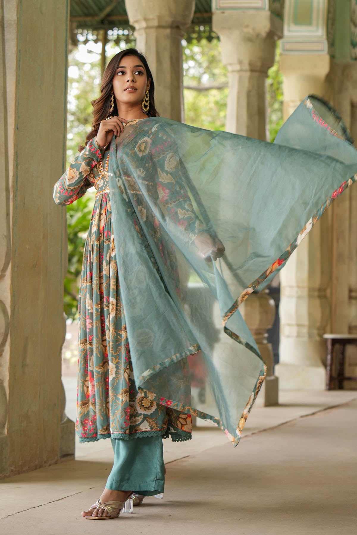 Woman holding a floral dupatta in an outdoor setting with architectural columns.