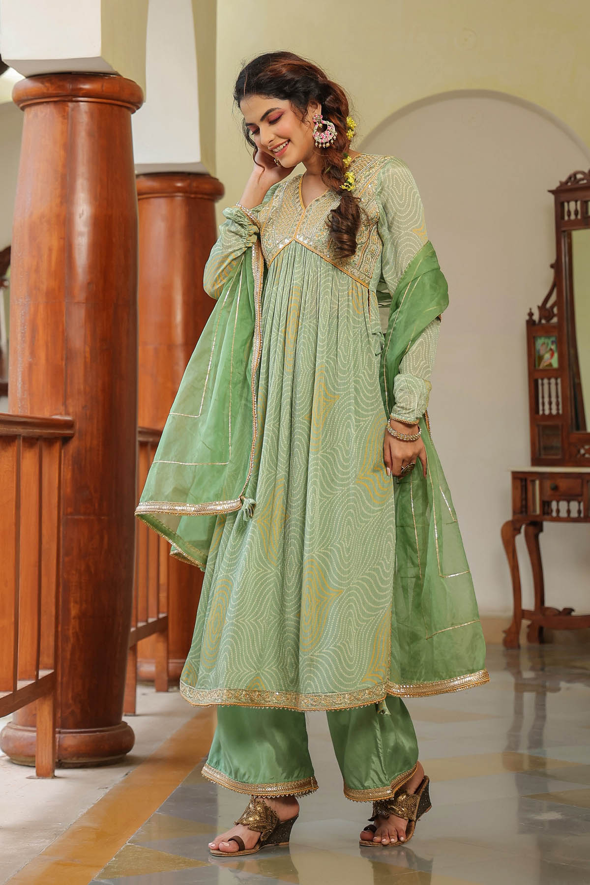 Woman in a green traditional outfit standing indoors with wooden columns and furniture.