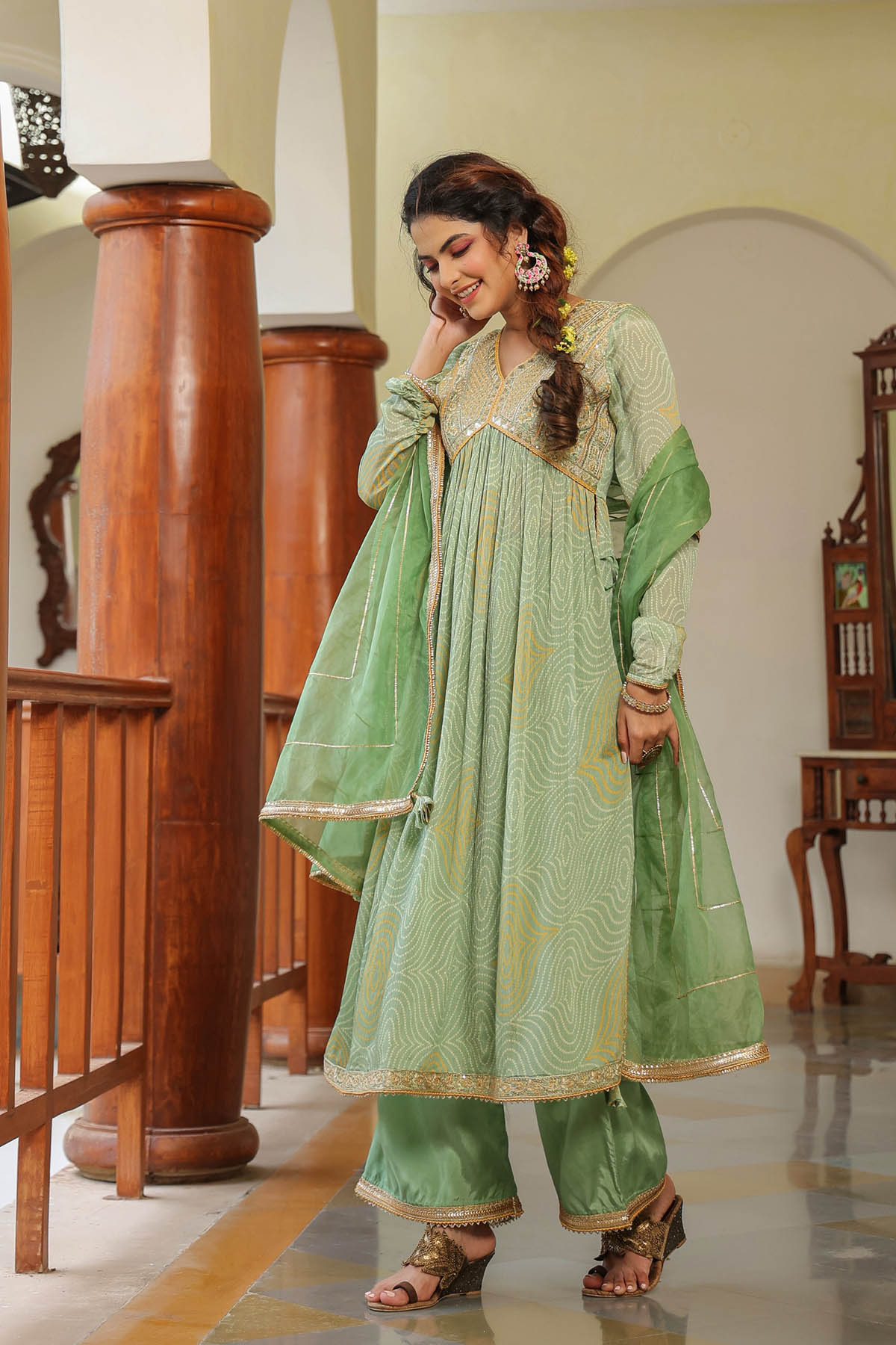 Woman in a green traditional outfit standing in a well-lit room with wooden columns.