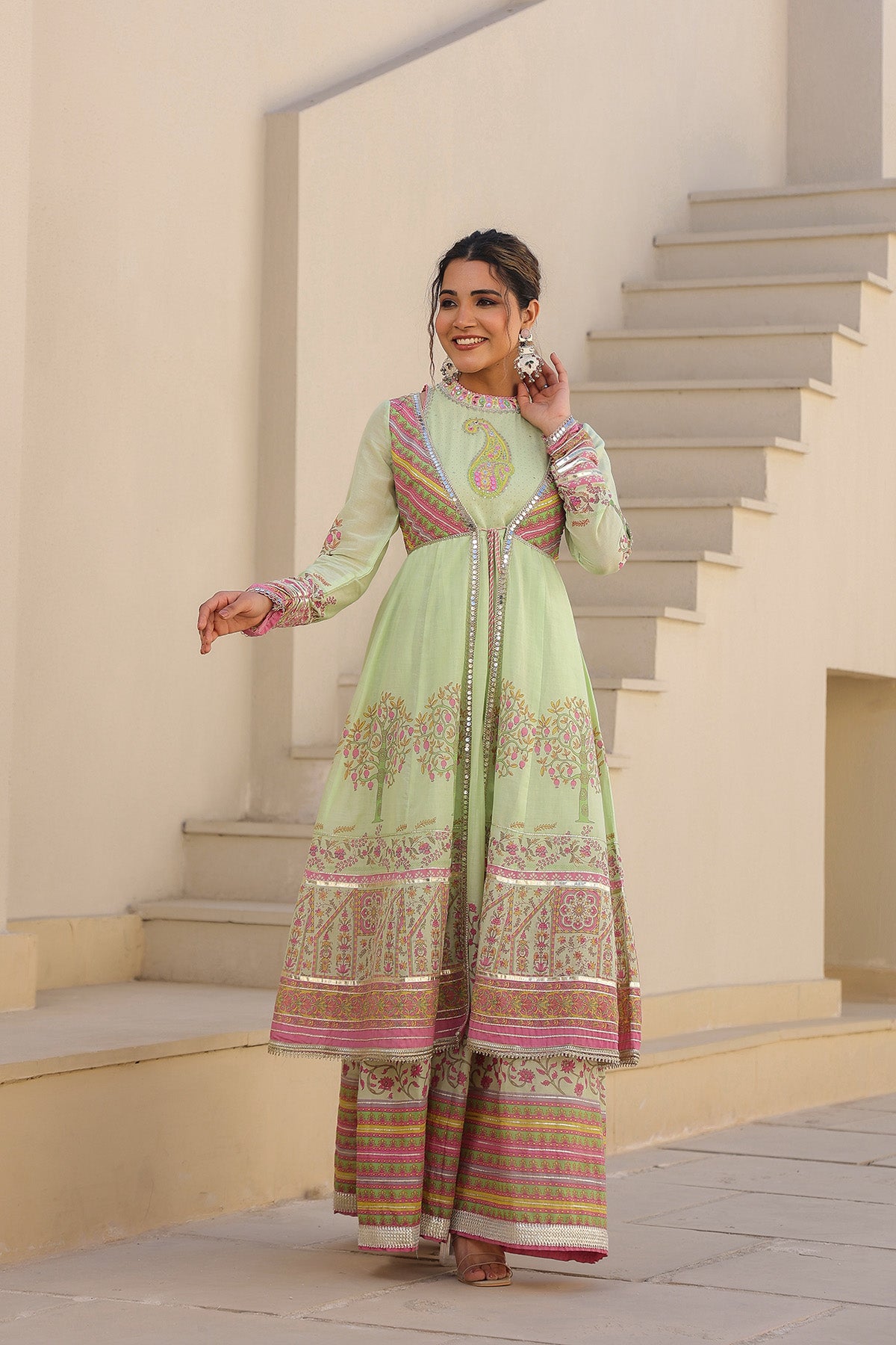 Woman in a light green traditional outfit with pink and gold patterns standing on steps.