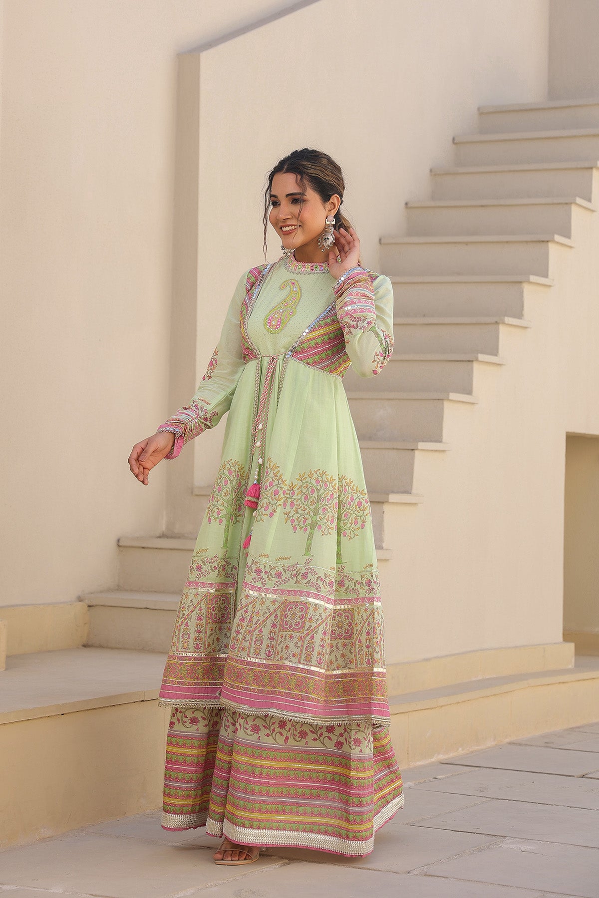 Woman in a light green traditional outfit with pink and gold patterns standing on a staircase.