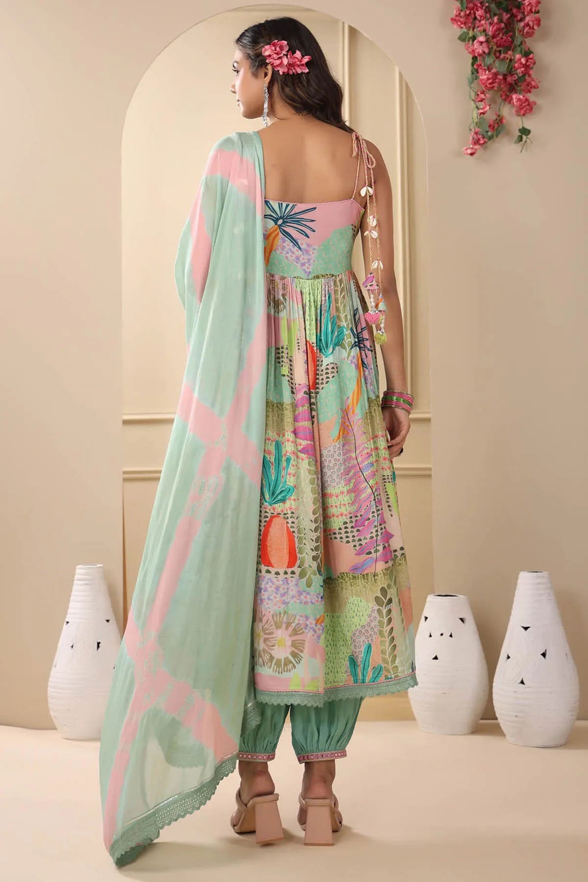 Woman in a colorful traditional outfit with a light green dupatta, standing in a room with decorative vases and flowers.