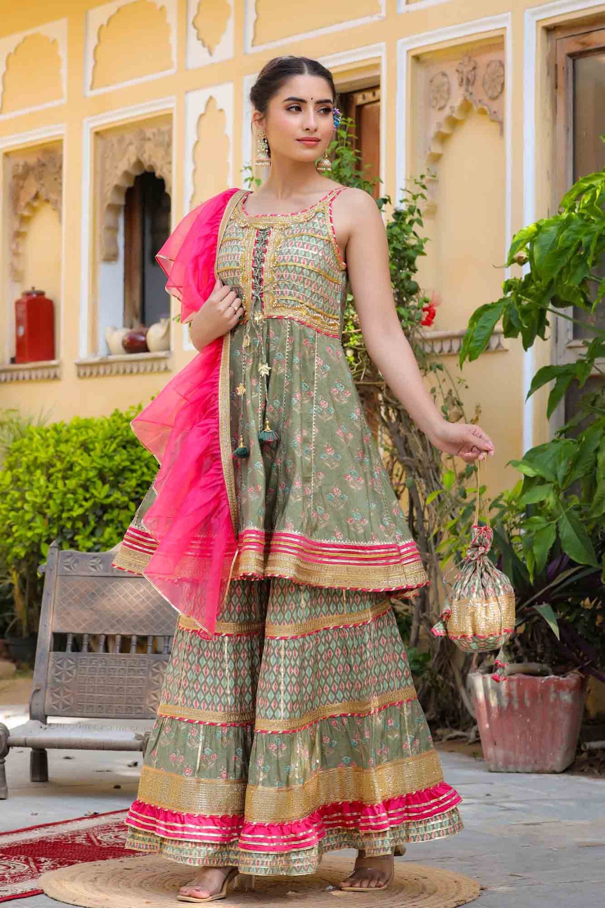 Woman in traditional outfit with pink dupatta standing in a decorative outdoor setting.