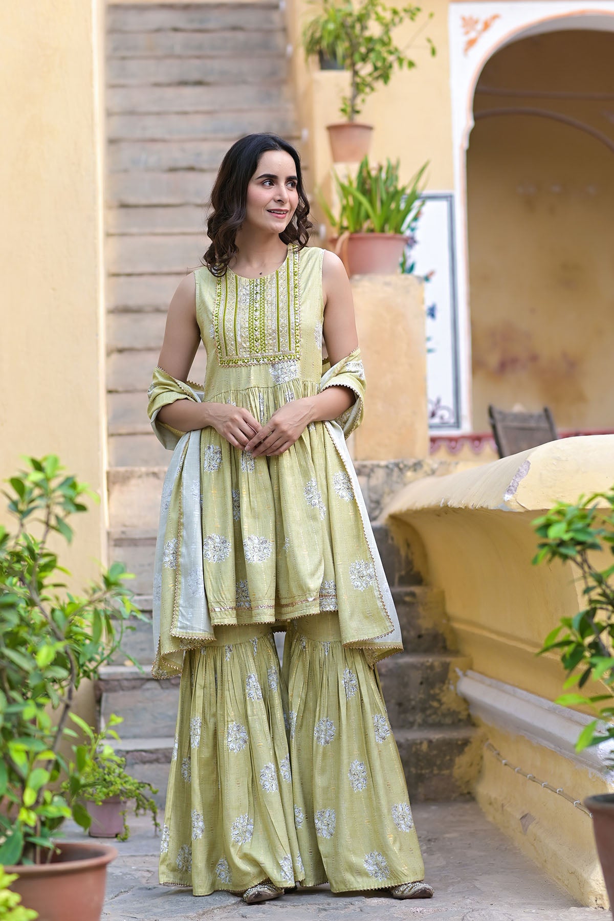 Woman in a green traditional outfit standing outdoors with plants and a yellow building in the background