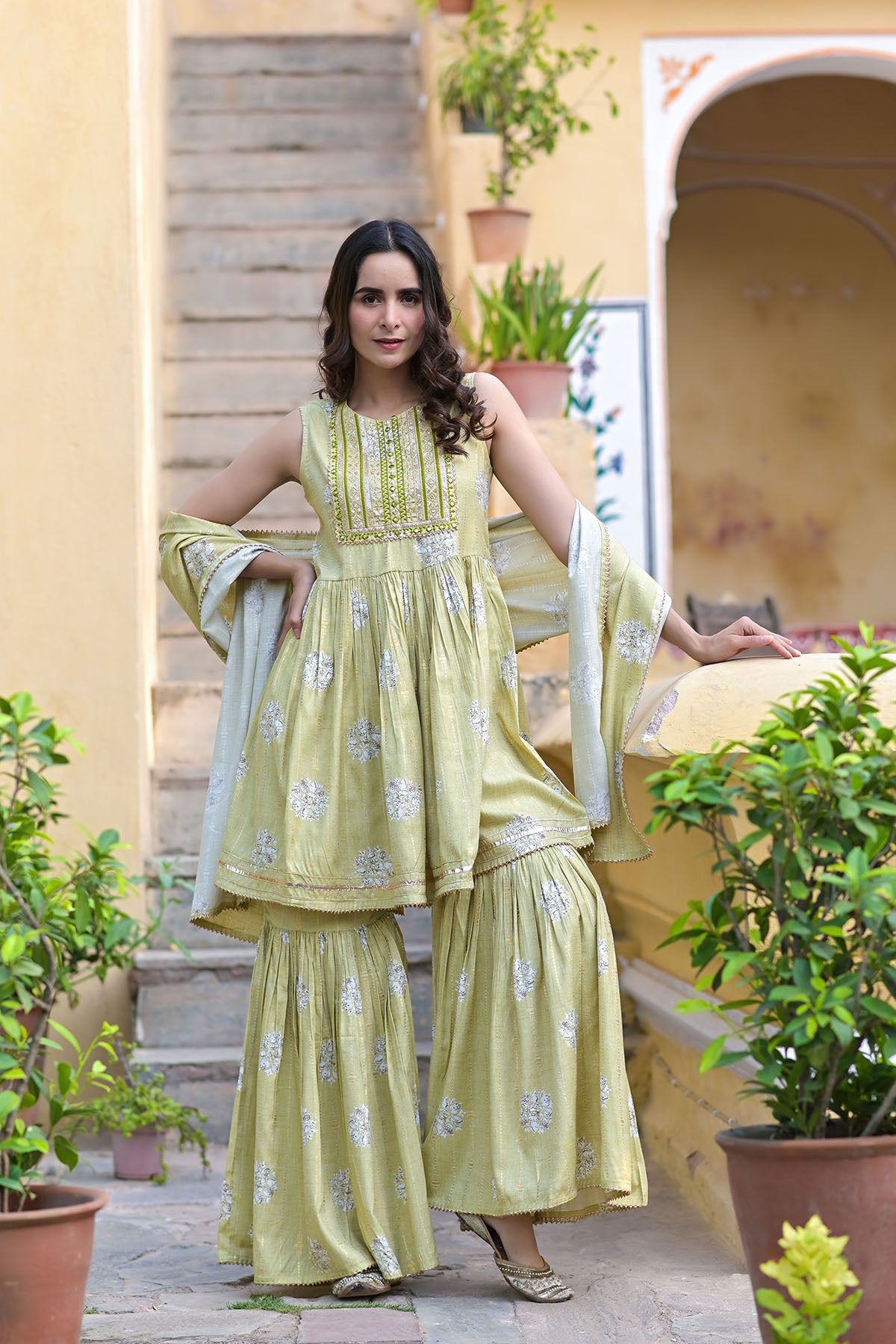 Woman in a light green traditional outfit standing on steps with plants around.