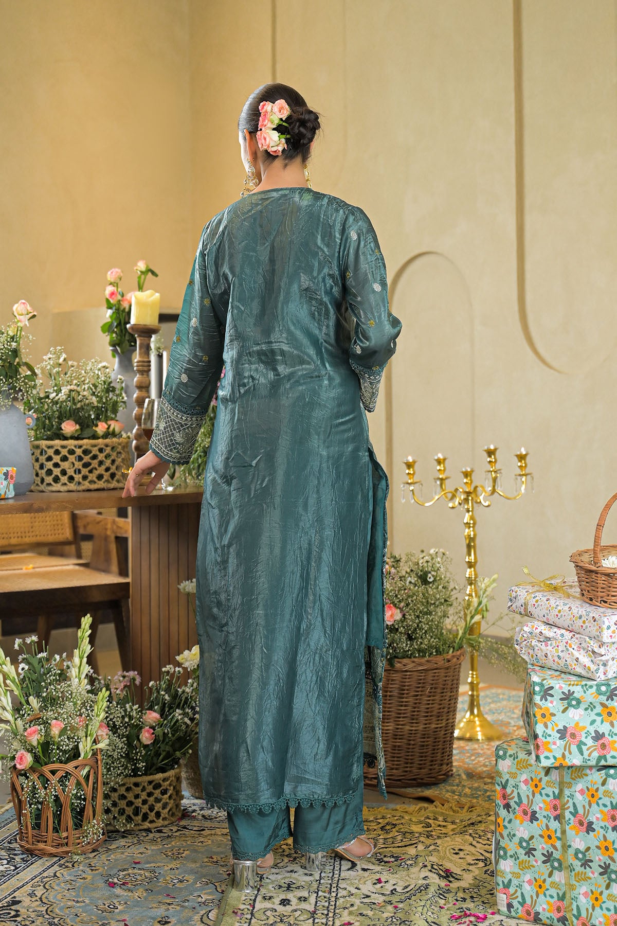Woman in a teal traditional outfit standing in a decorated room with flowers and a candelabra.
