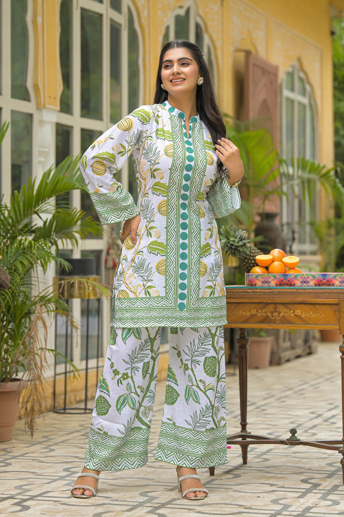 Woman in a white and green patterned outfit standing outdoors with plants and a table in the background.