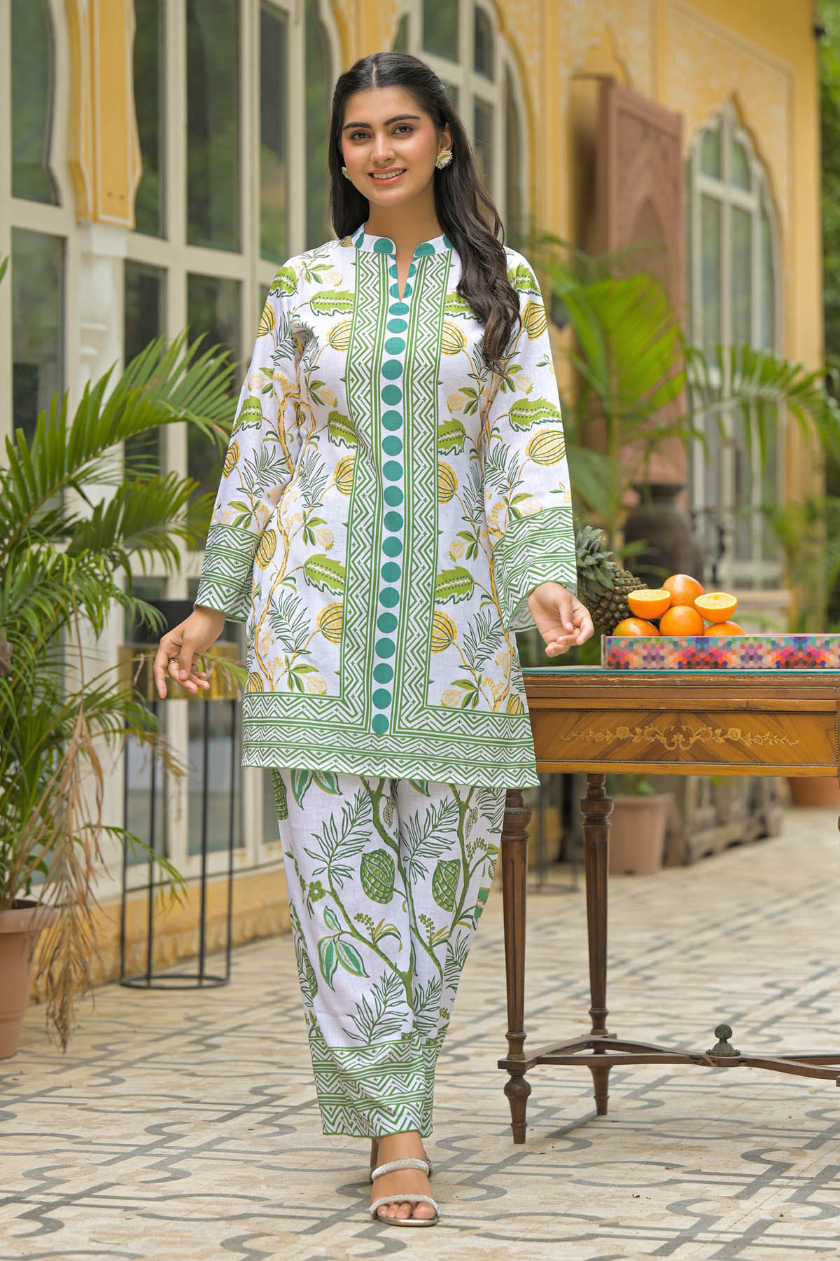 Woman in a green and white patterned outfit standing outdoors with plants and a table in the background.