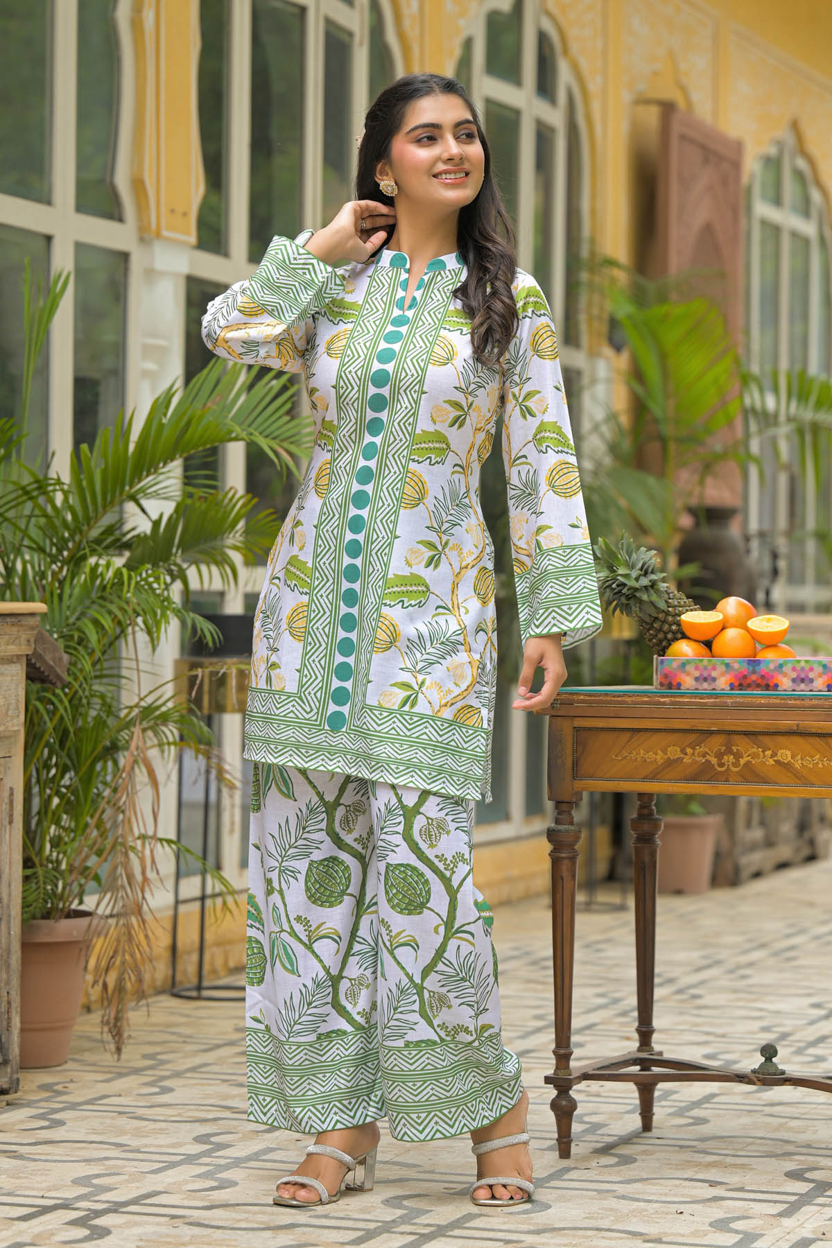 Woman in a green and white patterned outfit standing outdoors with plants and a table in the background.
