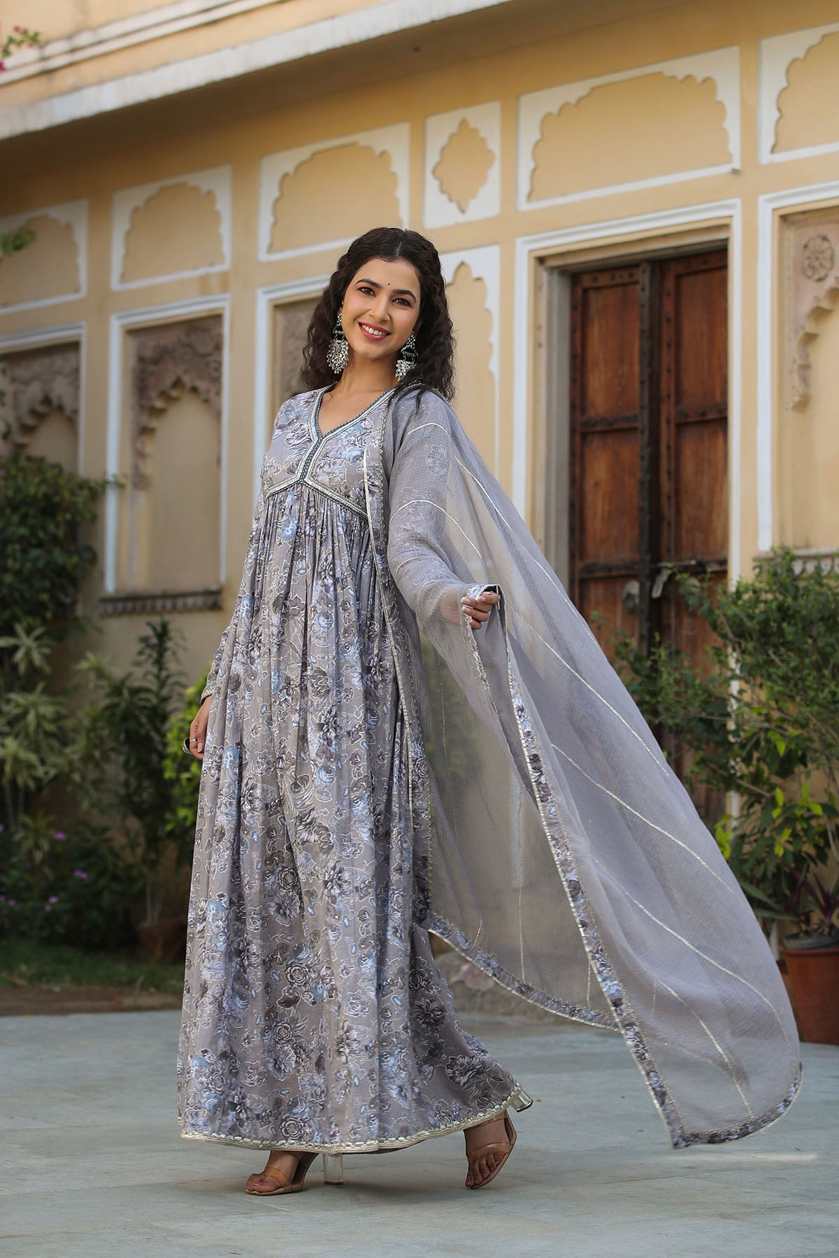 Woman in a floral dress with a sheer dupatta standing in front of a traditional building.