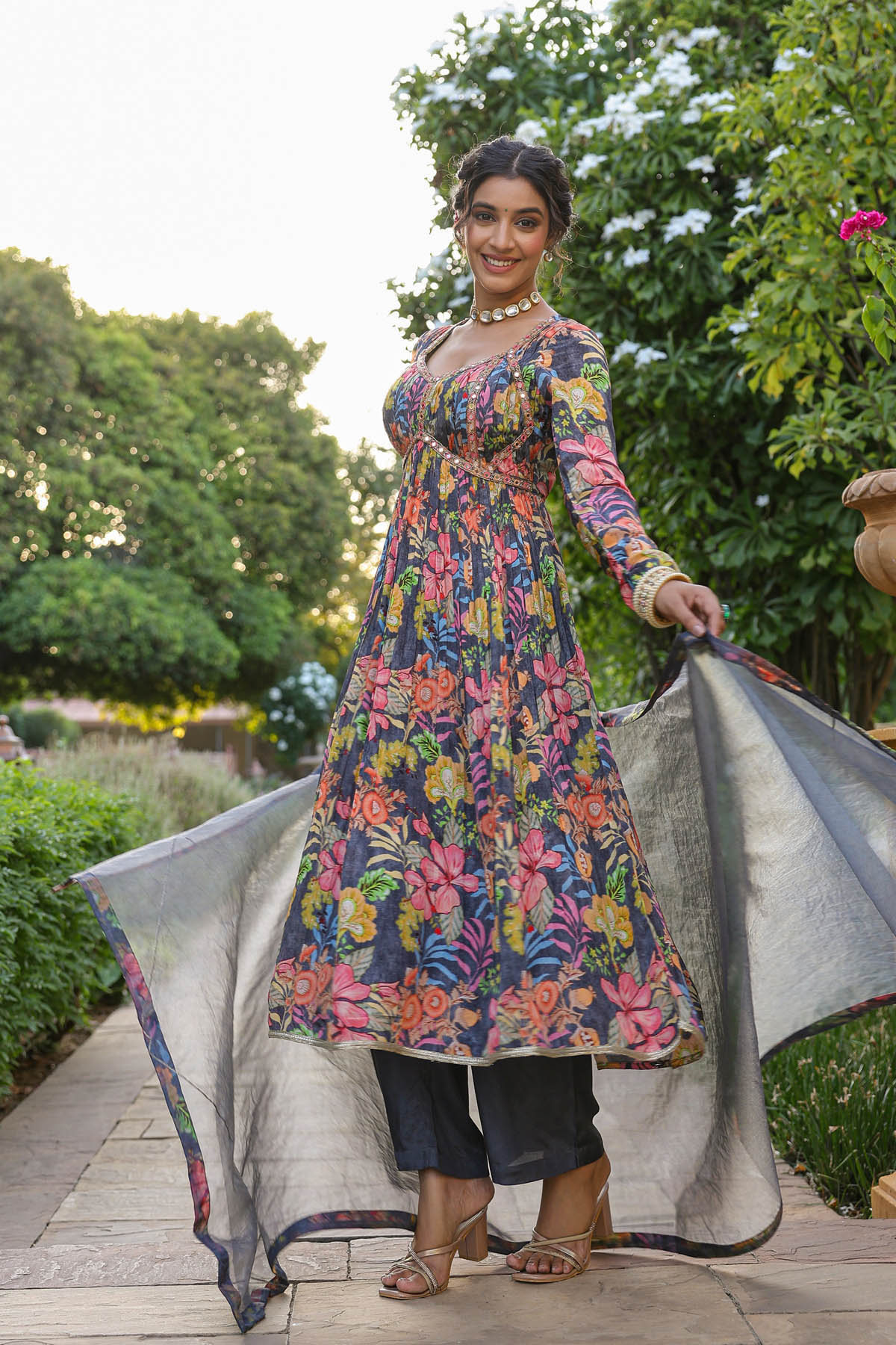 Woman in a colorful floral dress holding a silver dupatta in an outdoor setting.