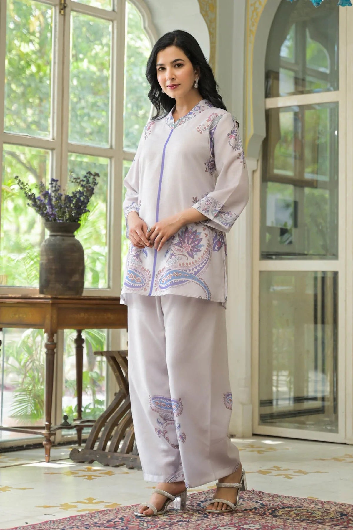 Woman in a light-colored outfit with floral patterns standing in a room with large windows and a table with plants.