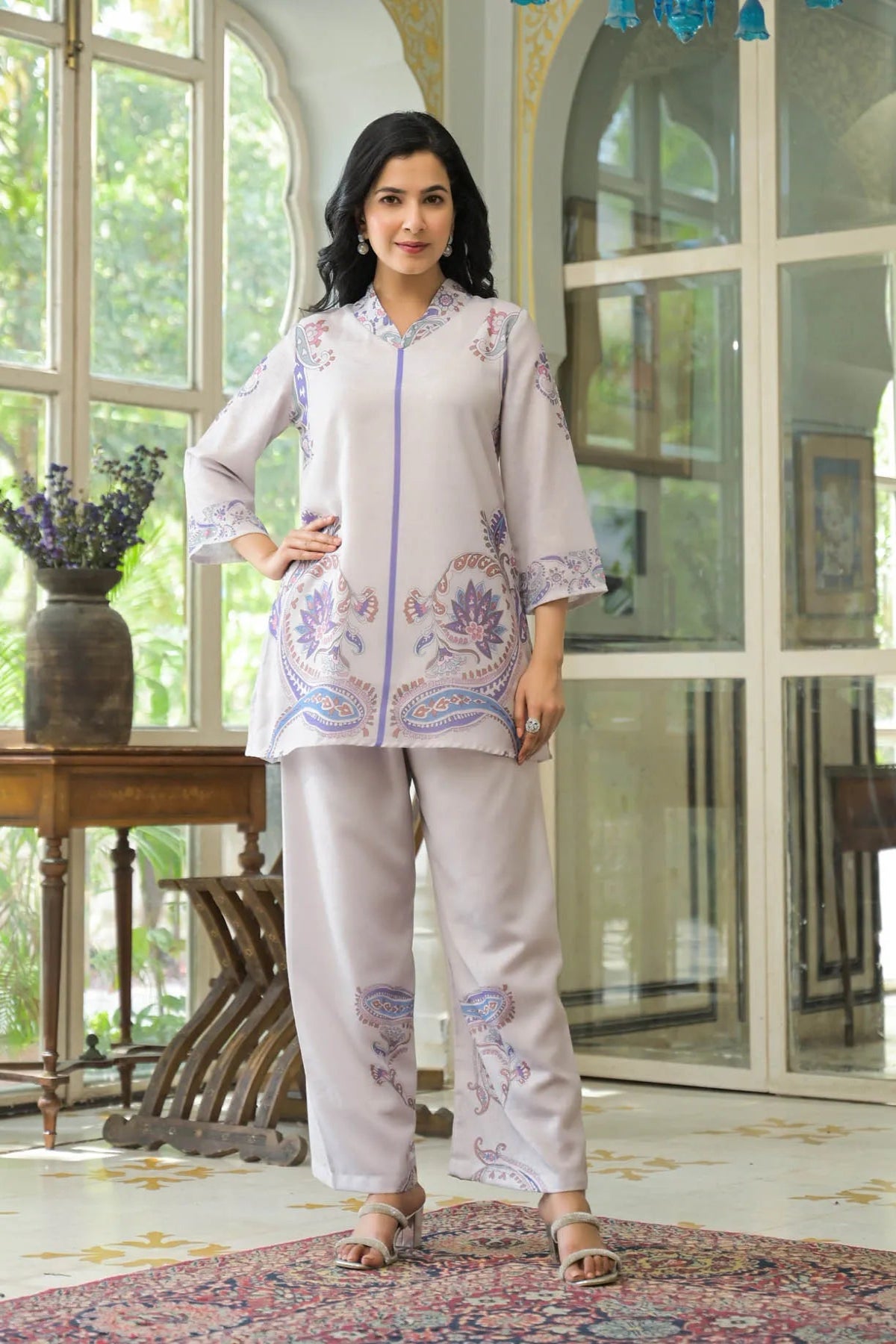 Woman wearing a light-colored embroidered outfit in a room with large windows.