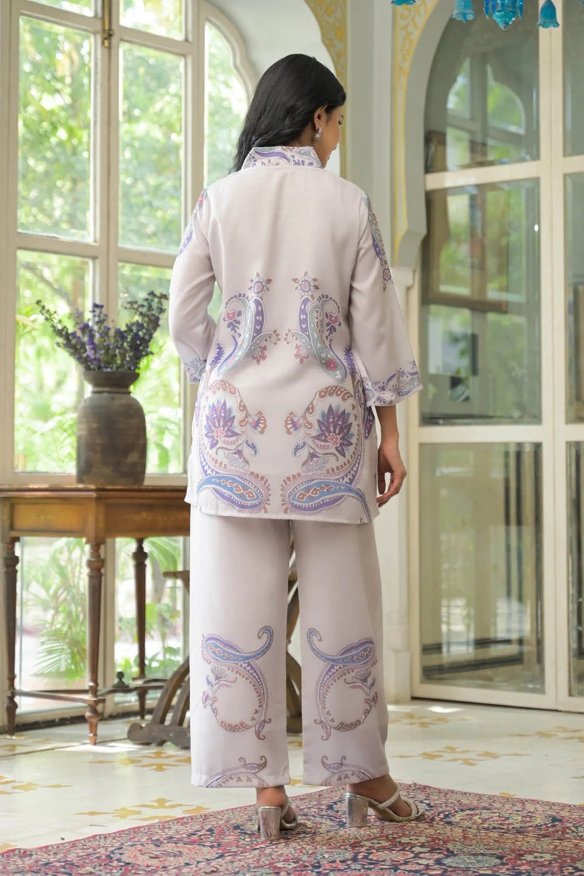 Woman wearing a cream-colored embroidered outfit standing in a room with large windows.