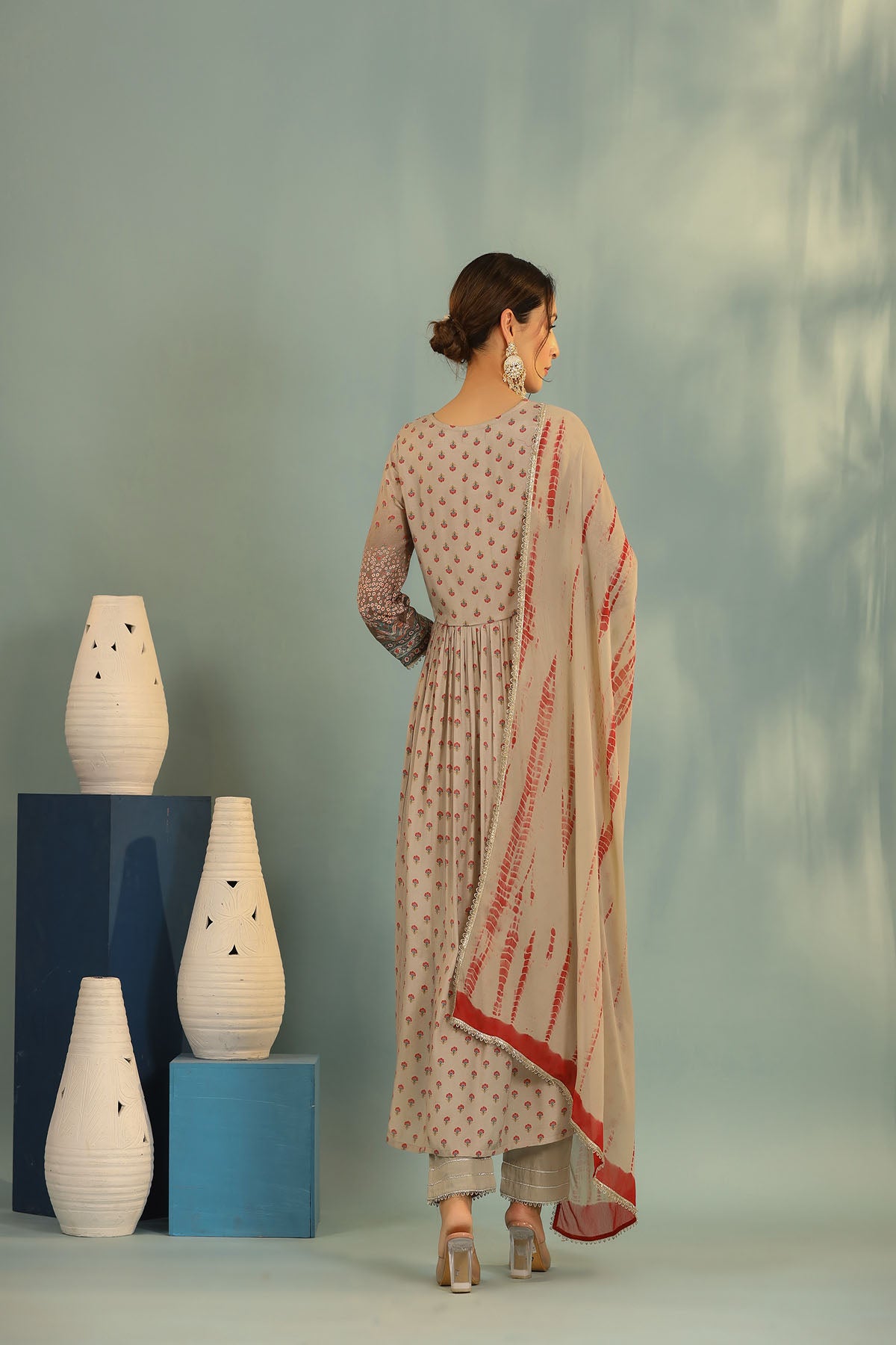 Woman in traditional outfit with a dupatta, standing next to decorative vases against a light green background