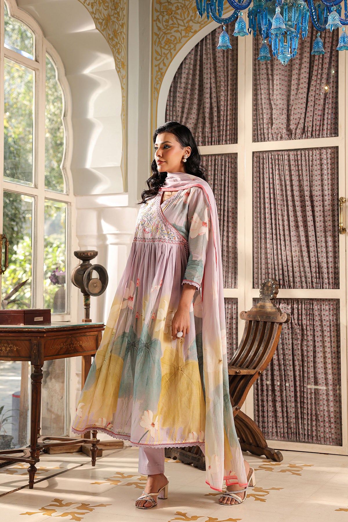 Woman in a traditional outfit standing in a decorated room with large windows and a chandelier.