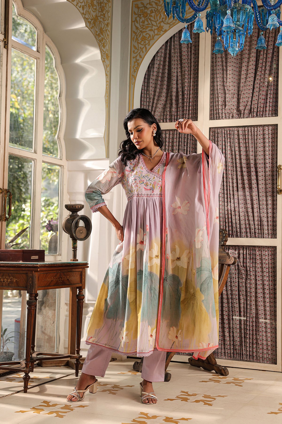 Woman in a colorful traditional outfit standing in an elegant room with large windows and decorative chandelier.