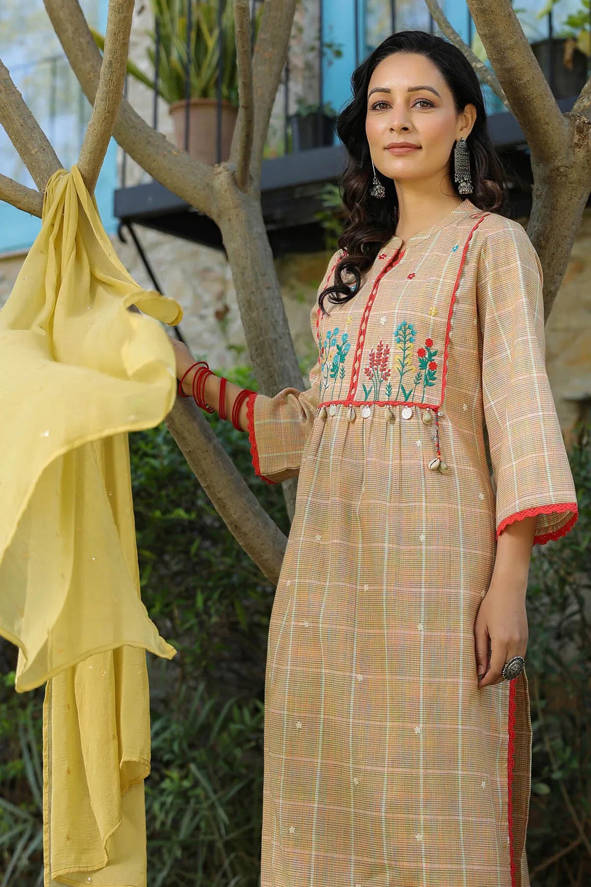 Woman wearing a traditional outfit with floral embroidery, standing outdoors near a tree.