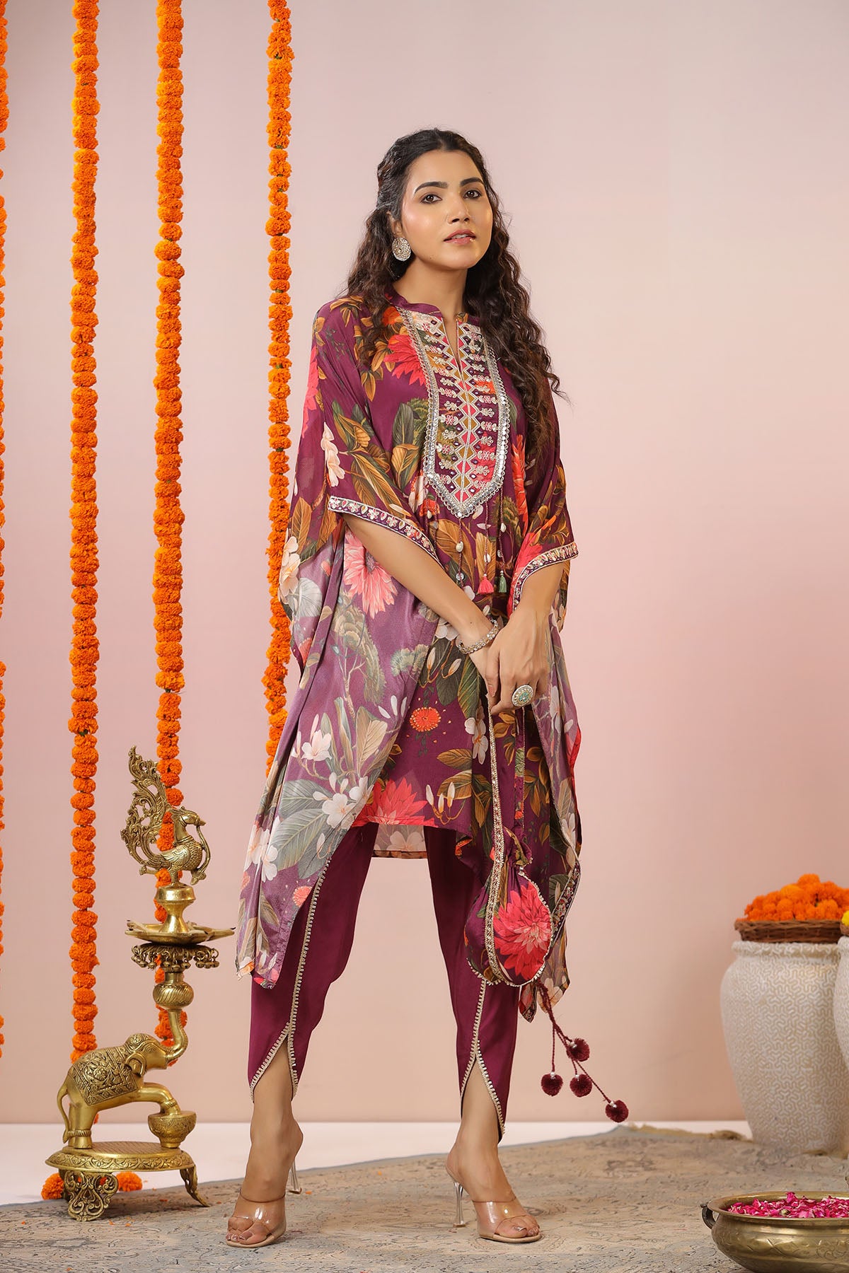 Woman in a colorful traditional outfit standing against a decorated background with orange flowers and a pink wall.