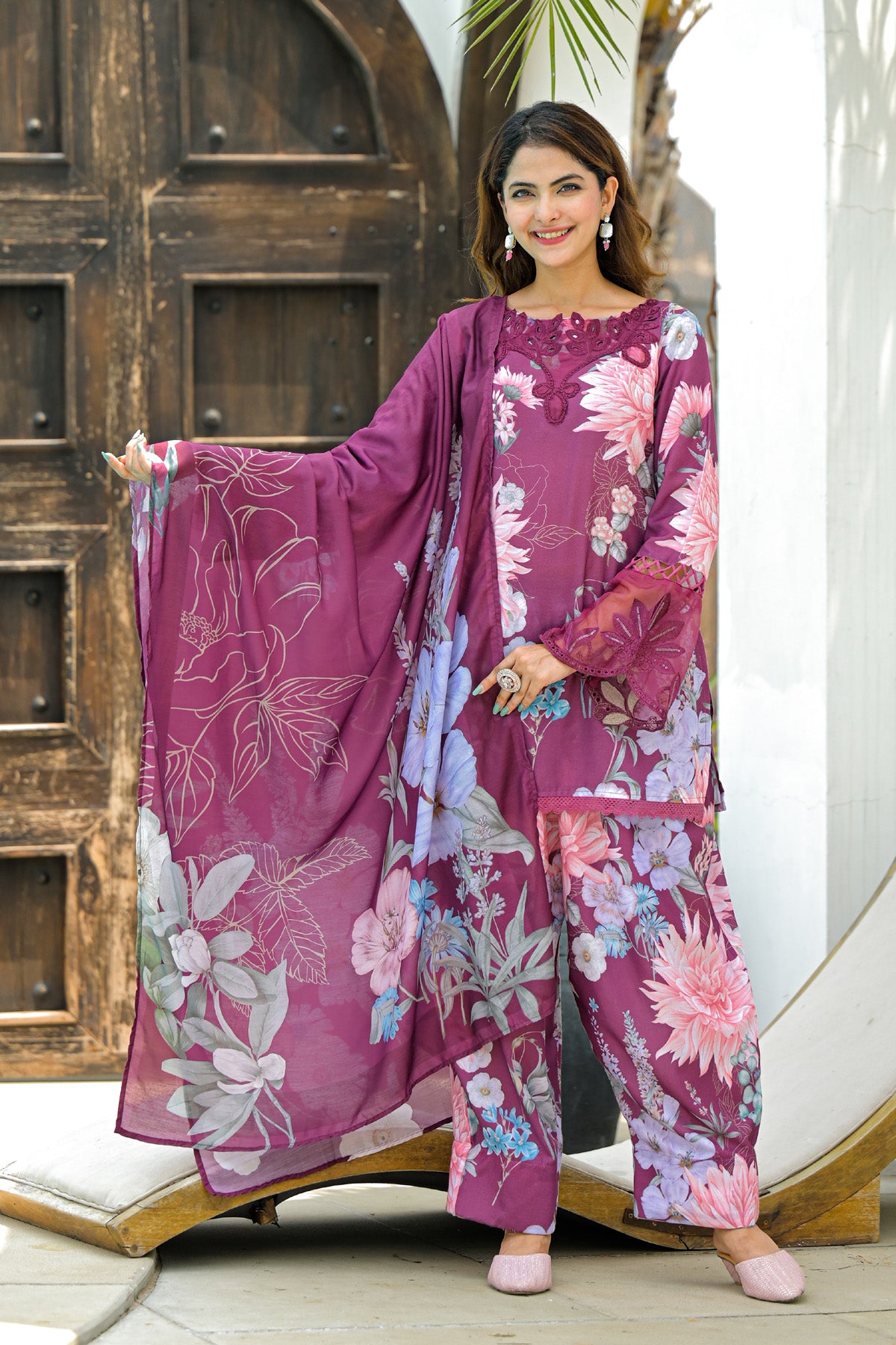 Woman wearing a floral purple outfit with a matching dupatta in front of a wooden door.