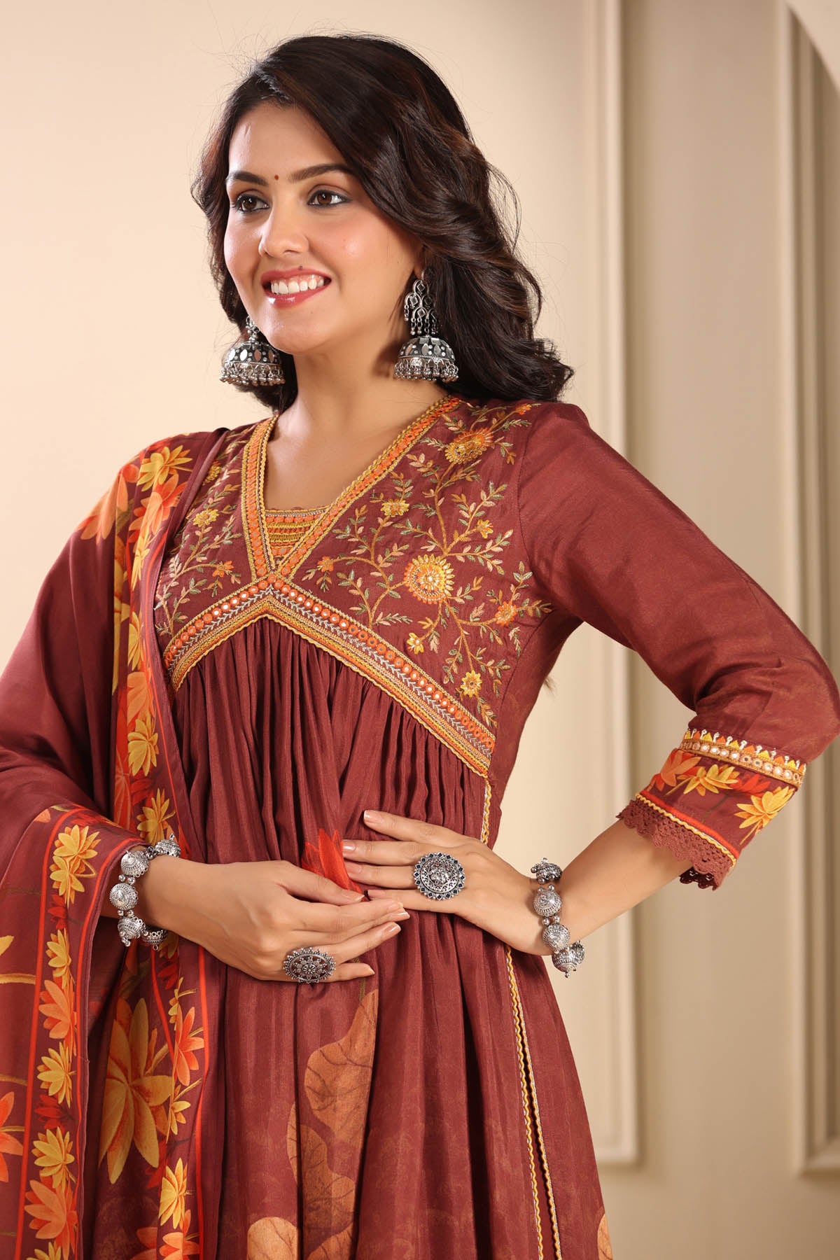 Woman wearing a traditional maroon and orange embroidered dress with jewelry against a beige wall.