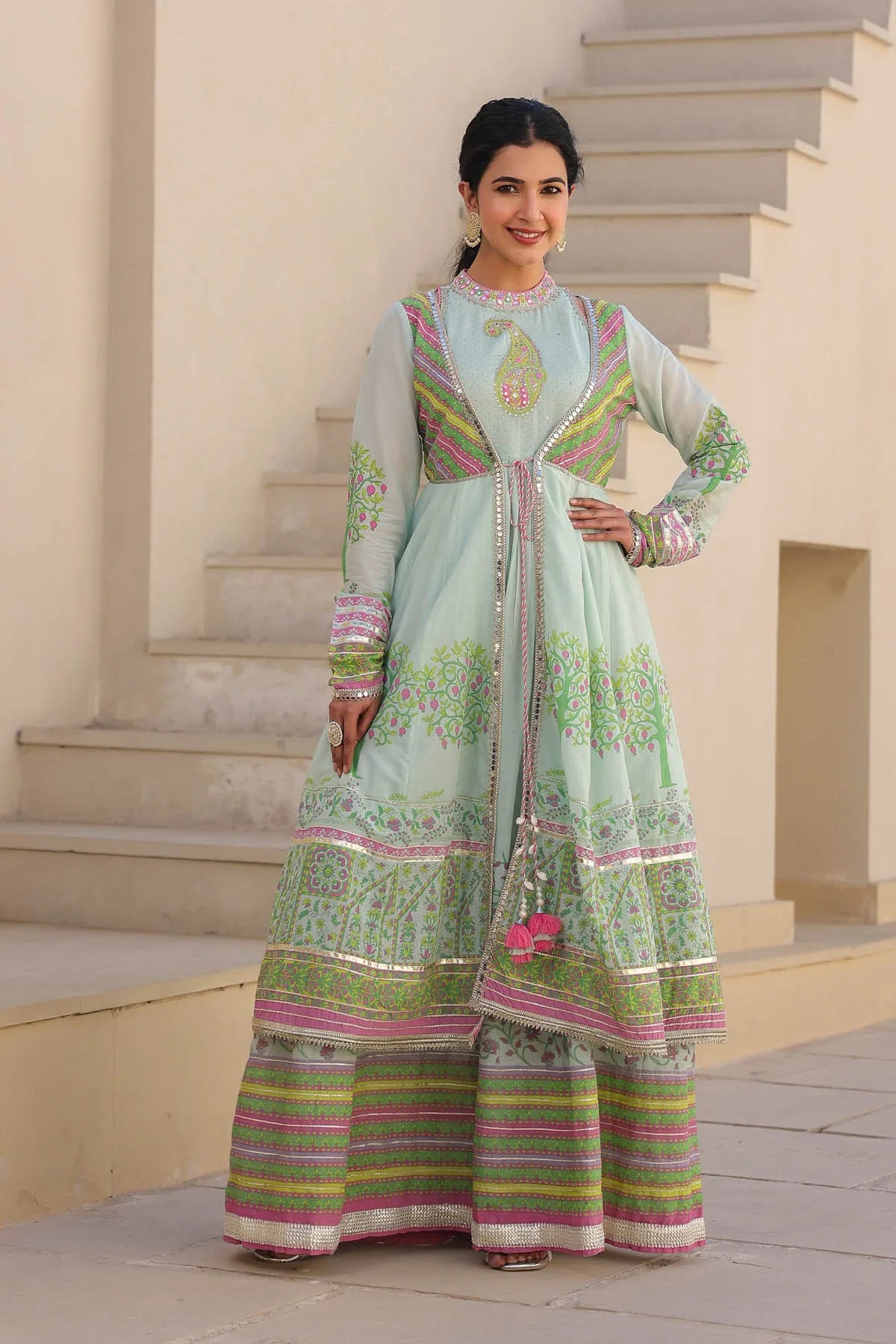 Woman wearing a light blue and green traditional outfit with floral patterns on a staircase.