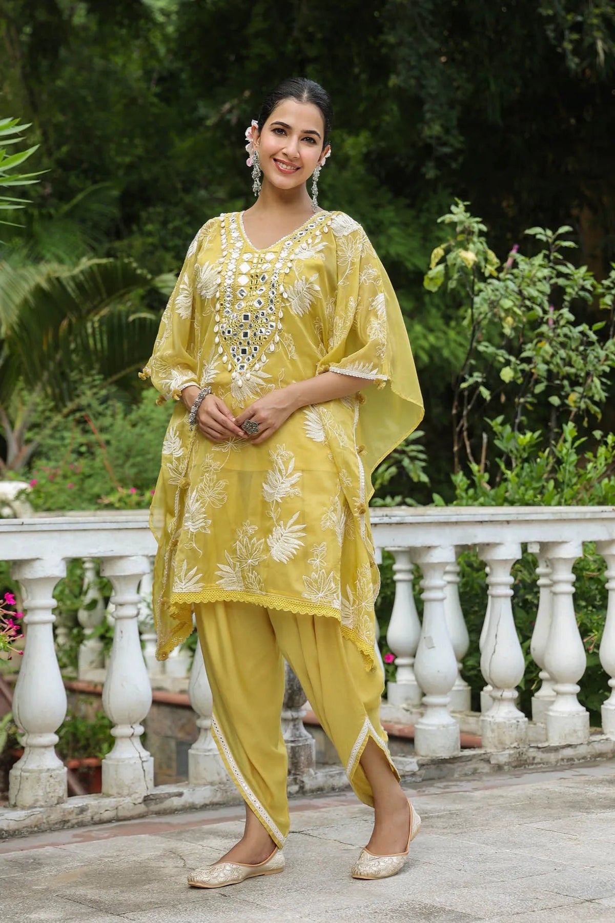 Woman in a yellow traditional outfit standing outdoors with greenery in the background