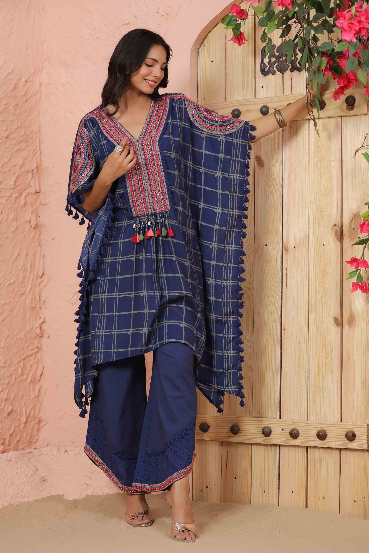 Woman wearing a blue checkered outfit with red patterns, standing against a wooden door with pink floral decorations.