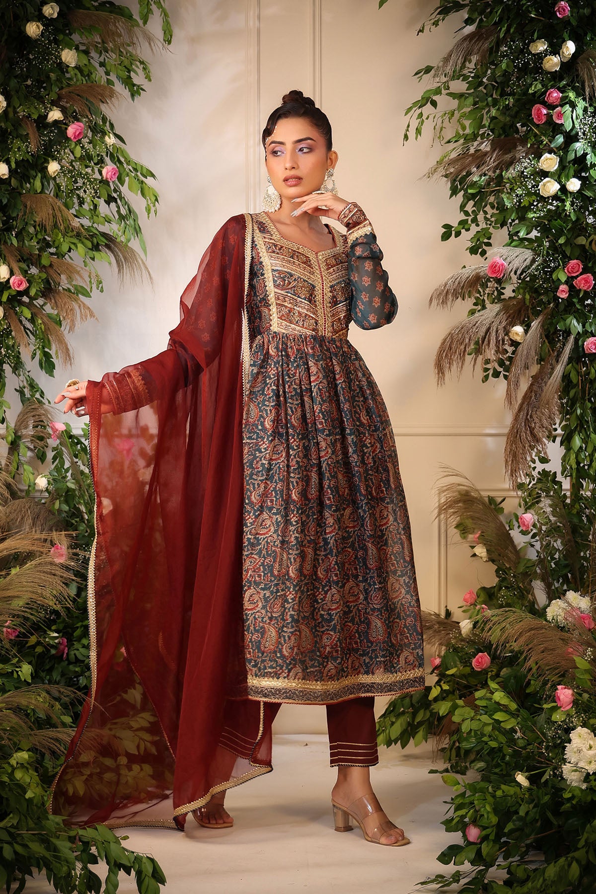 Woman in traditional outfit with floral patterns standing amidst decorative plants and flowers.