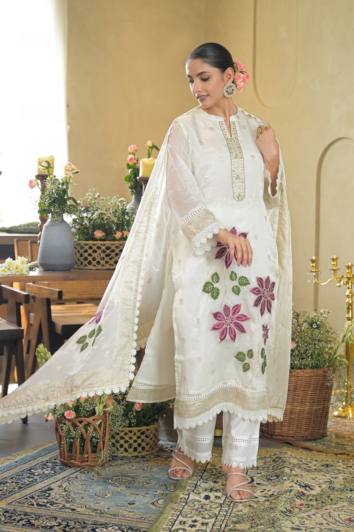 Woman in a white embroidered outfit standing in a decorated room with flowers and furniture.