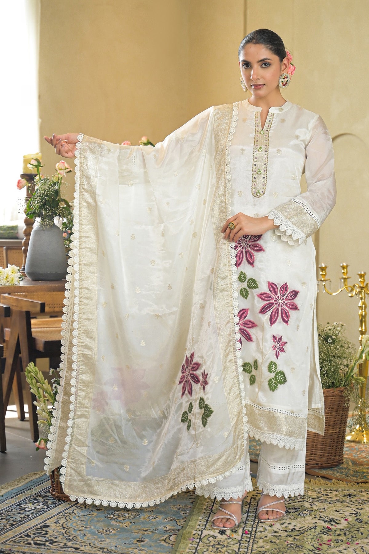 Woman in a white embroidered outfit with pink floral patterns in an elegant indoor setting.