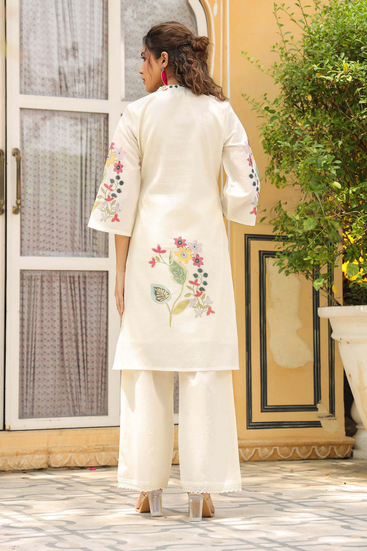 Woman wearing a white embroidered outfit standing in a decorative interior setting.