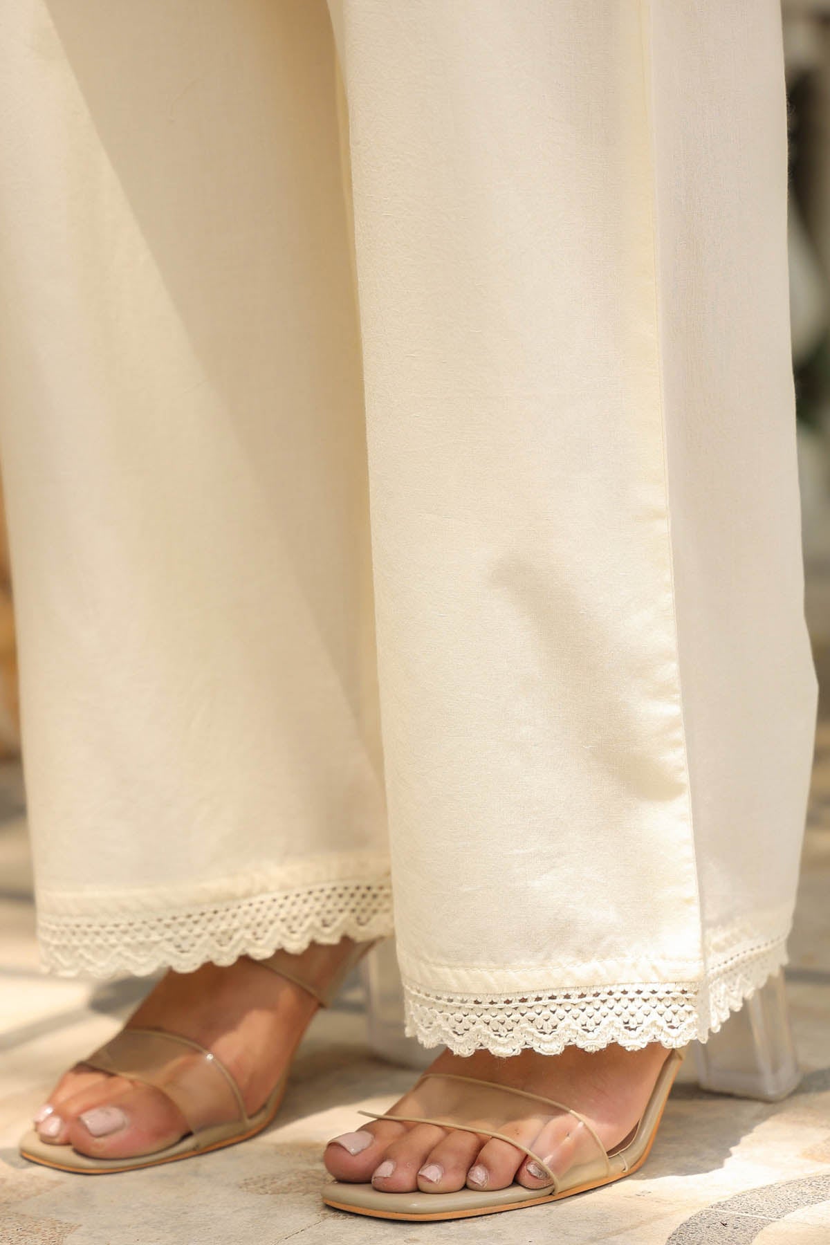 Close-up of white pants with lace trim and sandals on a neutral background
