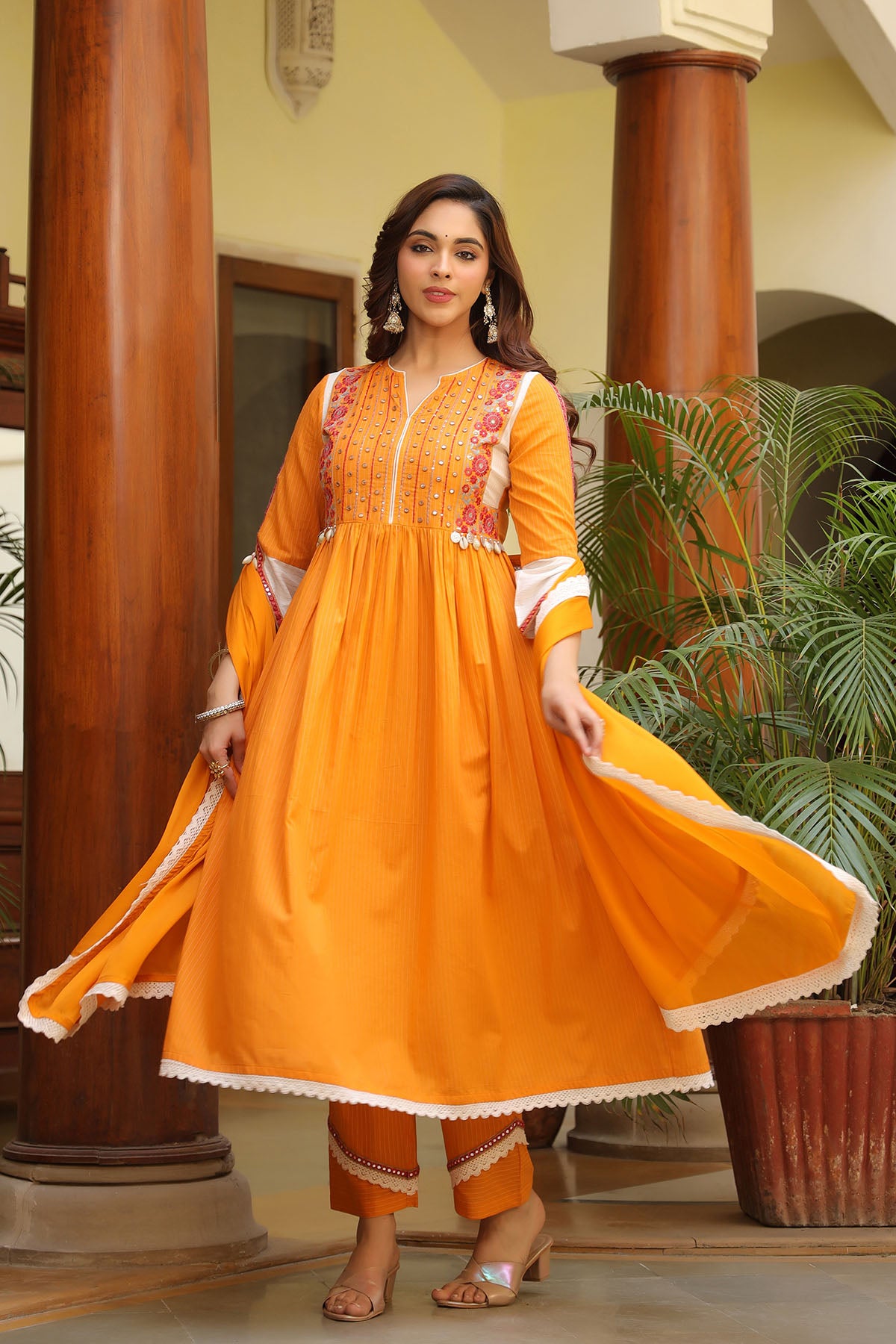 Woman in an orange traditional outfit standing in a decorative indoor setting with plants.