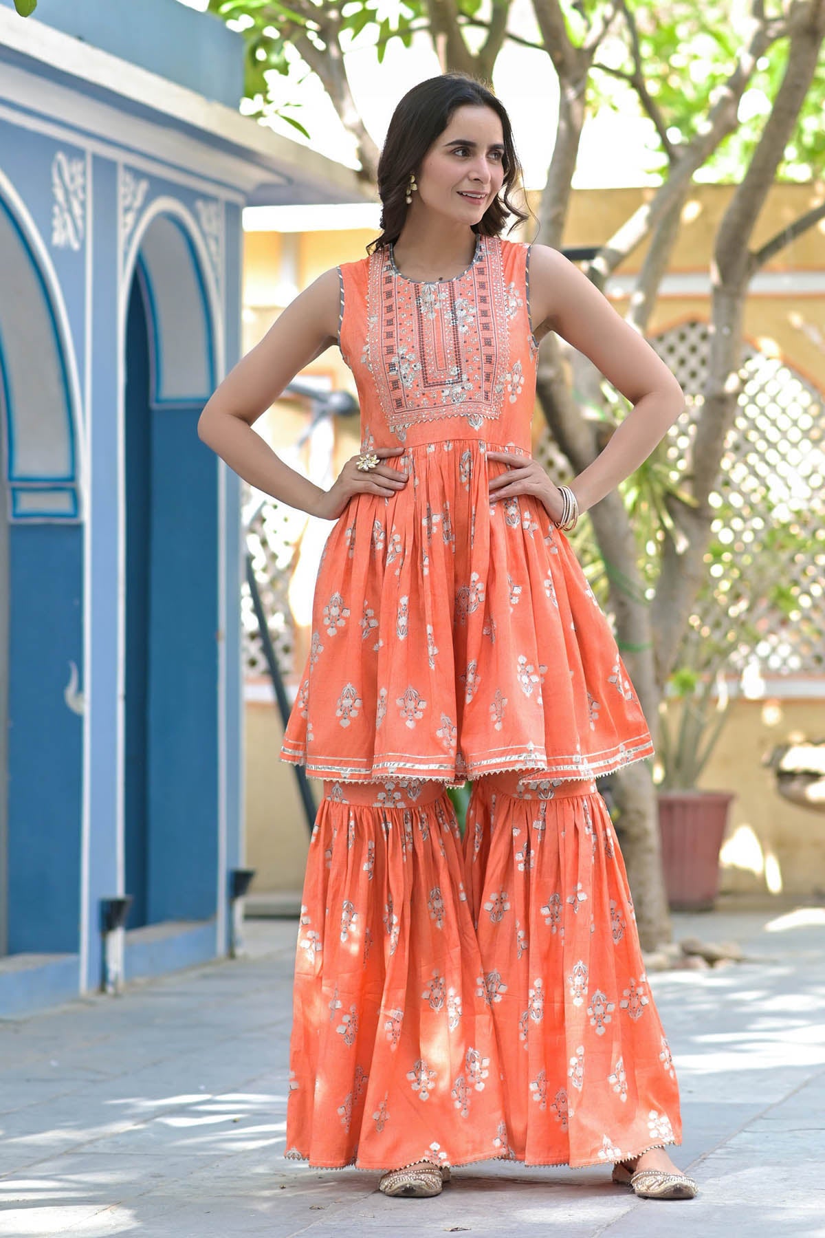 Woman wearing a coral floral dress standing outdoors with a blue archway and tree in the background.