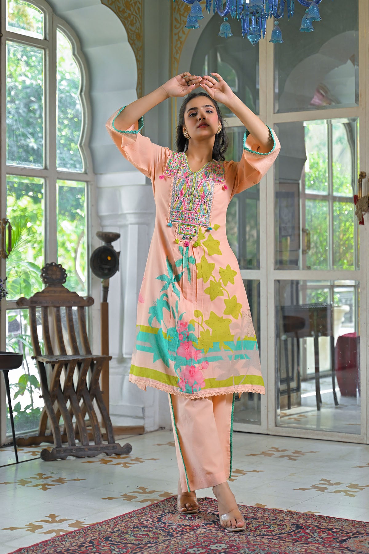 Woman in a colorful traditional outfit standing in a well-lit room with large windows.
