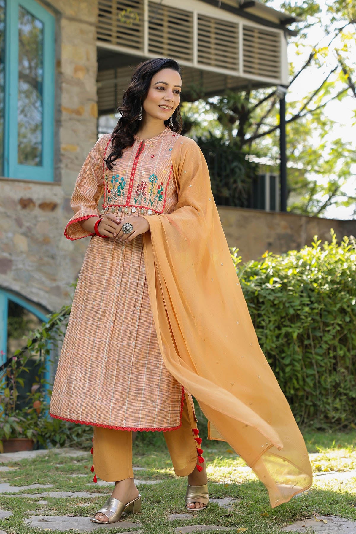 Woman in traditional outfit standing outdoors with a building and greenery in the background