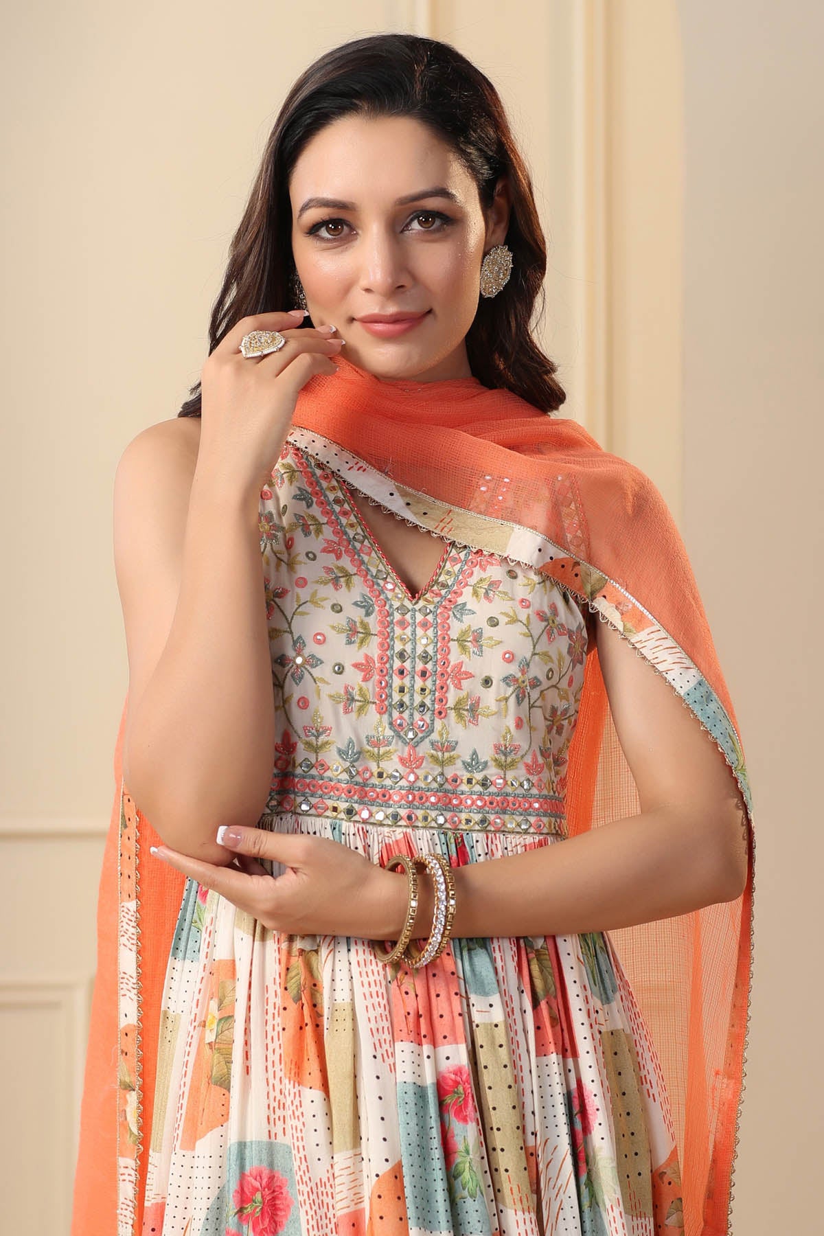 Woman wearing a patterned dress with an orange dupatta against a beige background