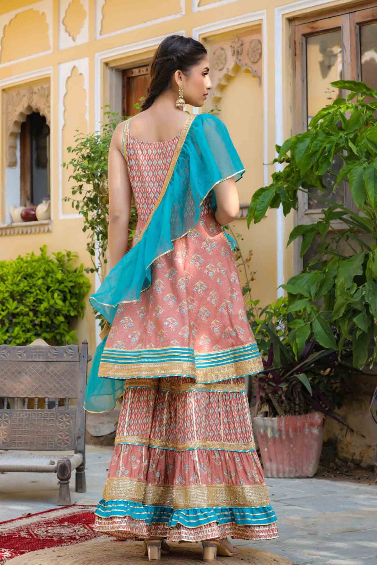 Woman in a traditional pink and blue saree standing outdoors with plants and a building in the background.