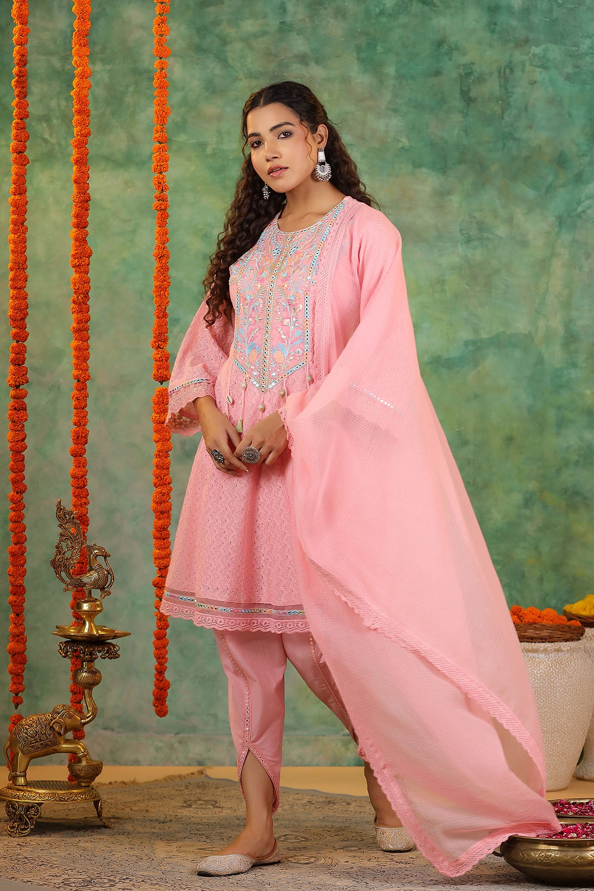 Woman in a pink traditional outfit standing against a decorative background with orange strings and green wall.