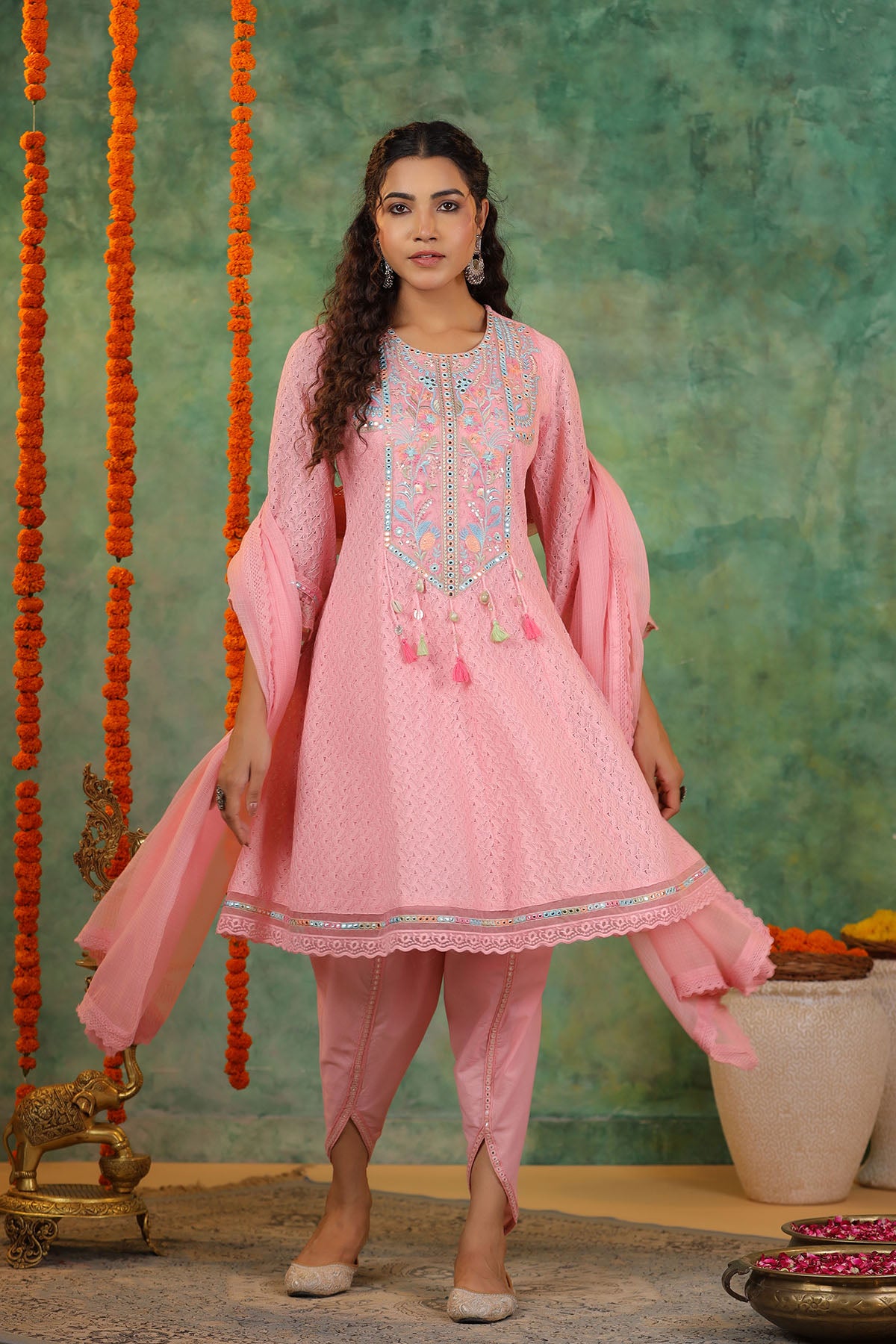 Woman in a pink traditional outfit with embroidery standing against a textured green wall.